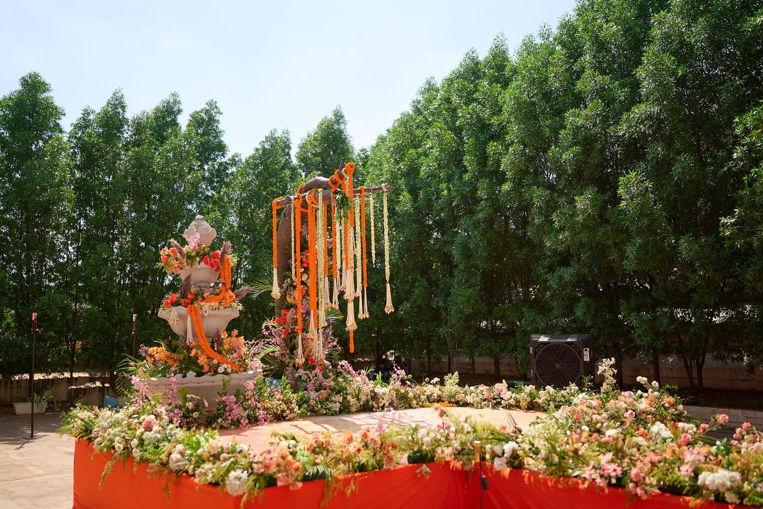 Vibrant Indian wedding mandap decorated with orange marigold garlands, colorful flowers, and a deity statue outdoors.