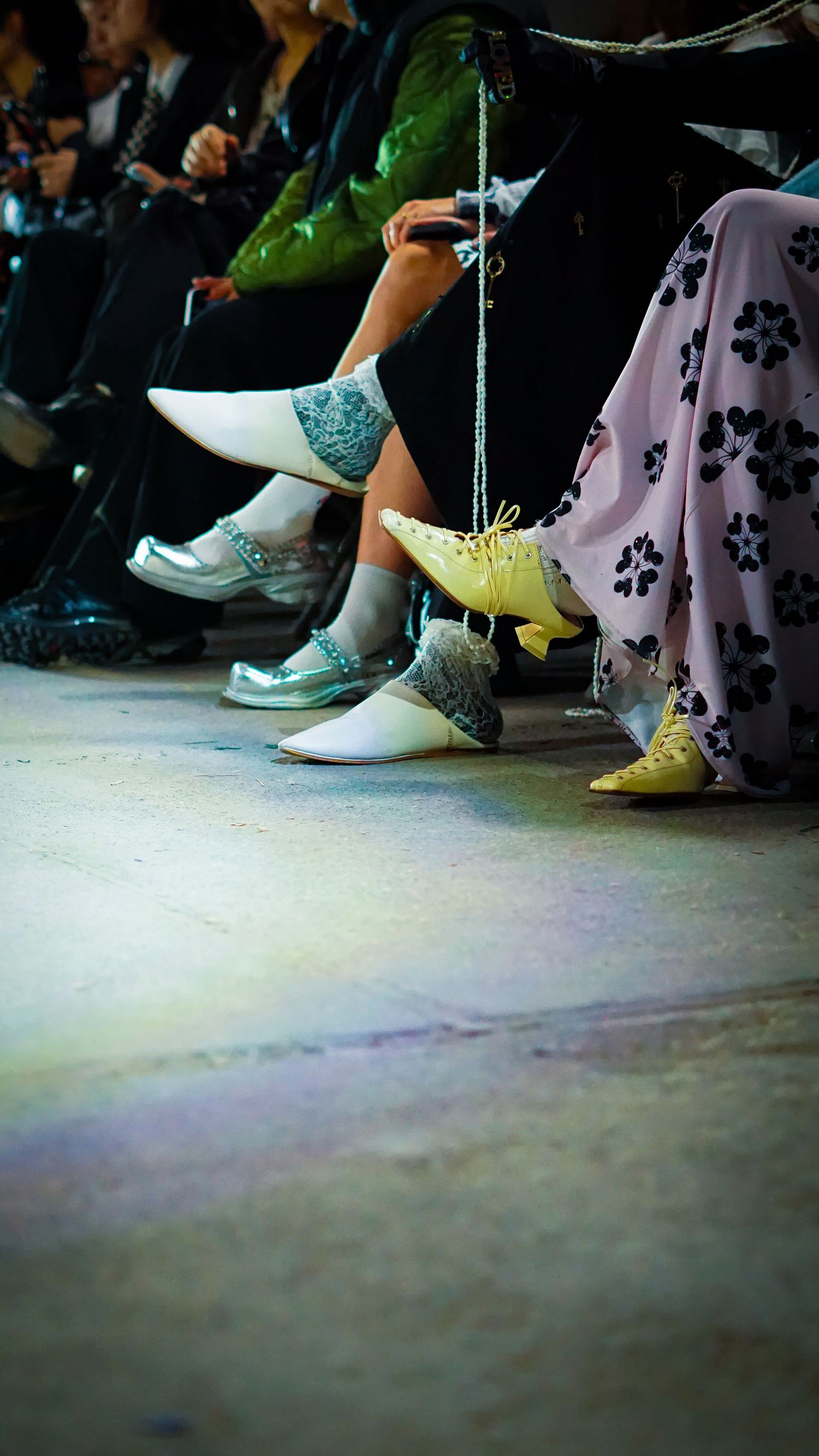 Close-up of fashion show audience feet wearing unique designer shoes including yellow lace-ups and silver pointed flats.