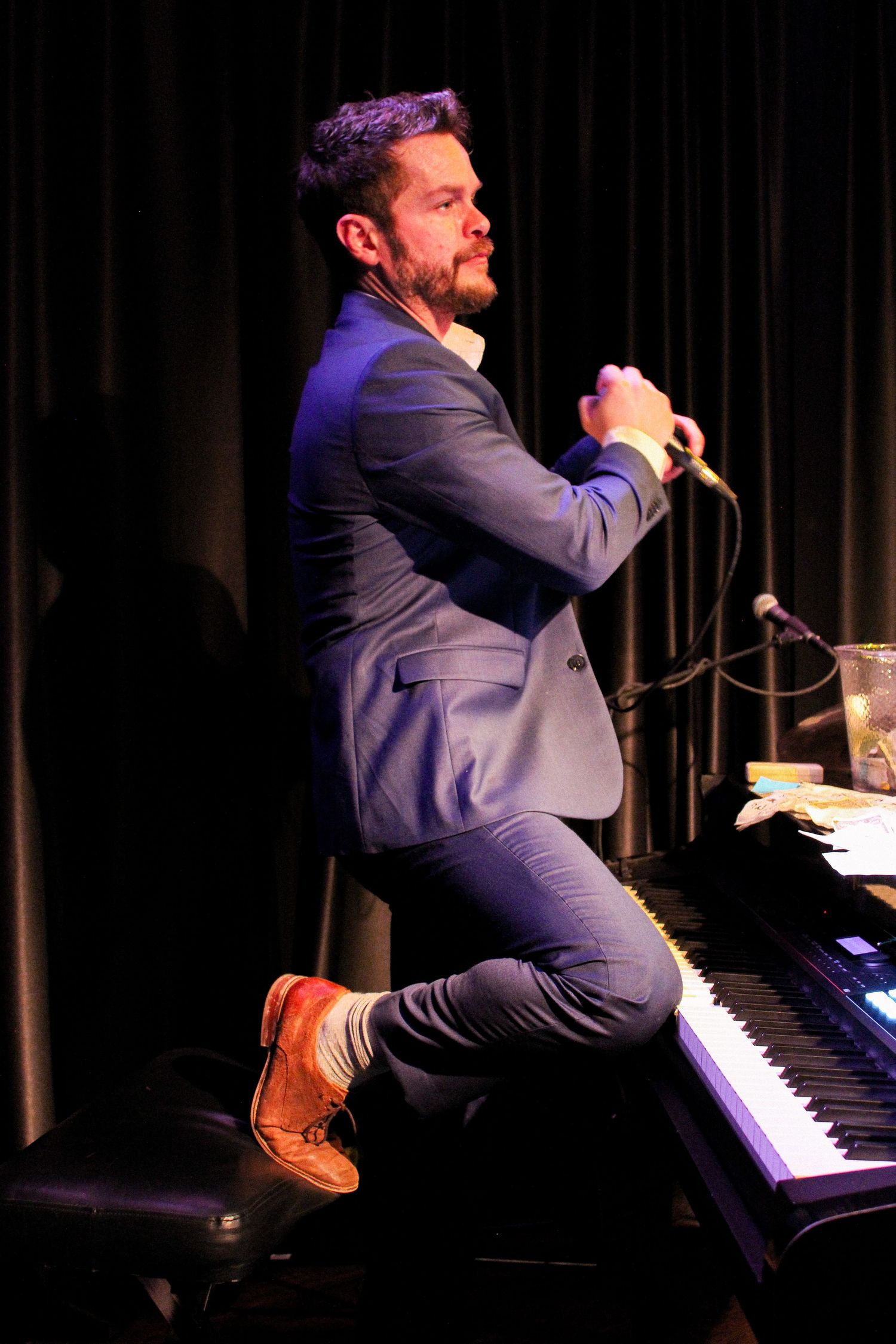 Bearded man in blue suit perched on piano bench holding microphone on stage.