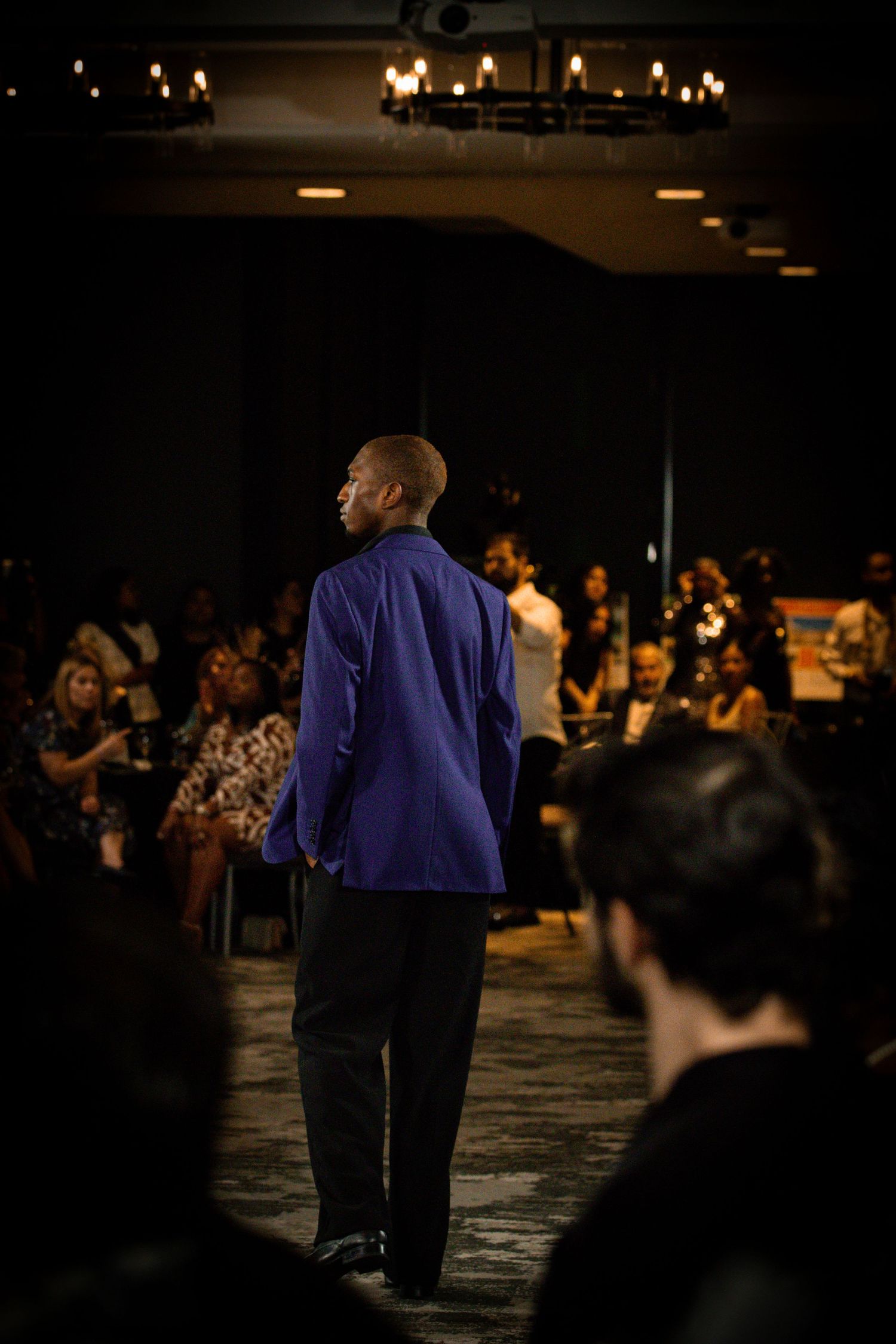 Man in blue blazer standing on a runway at an indoor fashion show with audience seated around him.