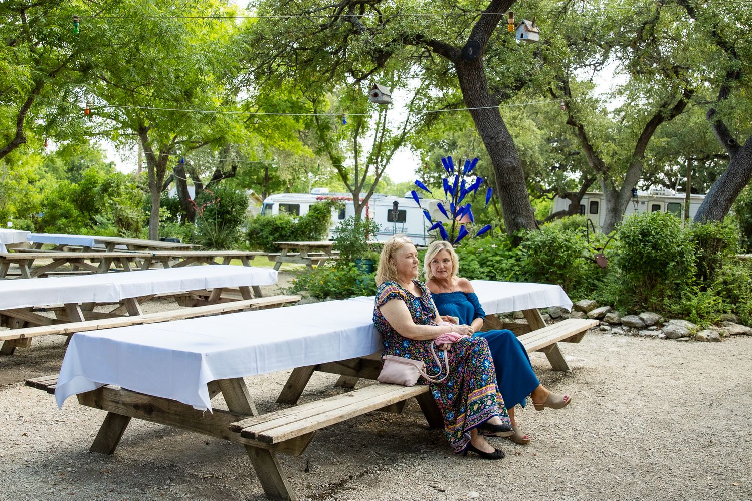 Two women sit together at a picnic table in a shaded outdoor garden area.