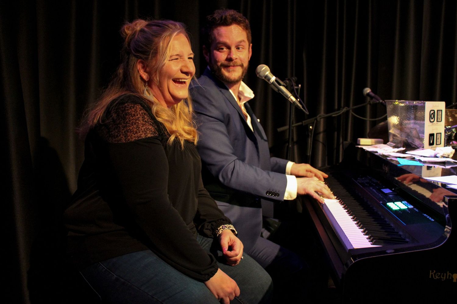 Smiling man and woman sitting together at a keyboard piano on stage.