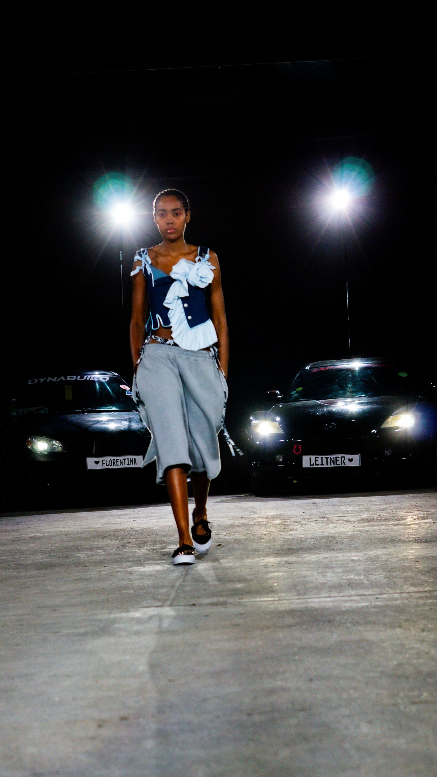 Model in denim and grey wide-leg pants walks fashion runway between two black luxury cars at night.
