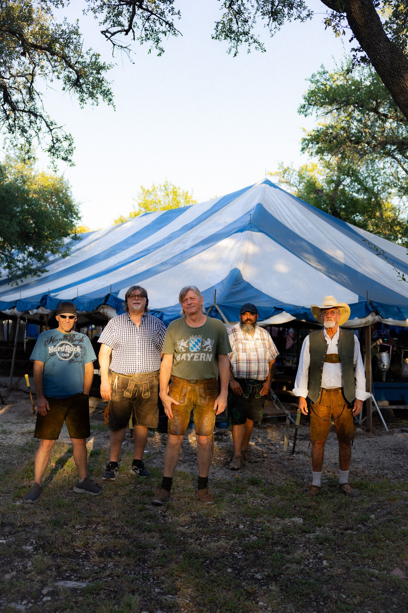 Five men in traditional German lederhosen standing together outdoors under a blue and white striped festival tent.