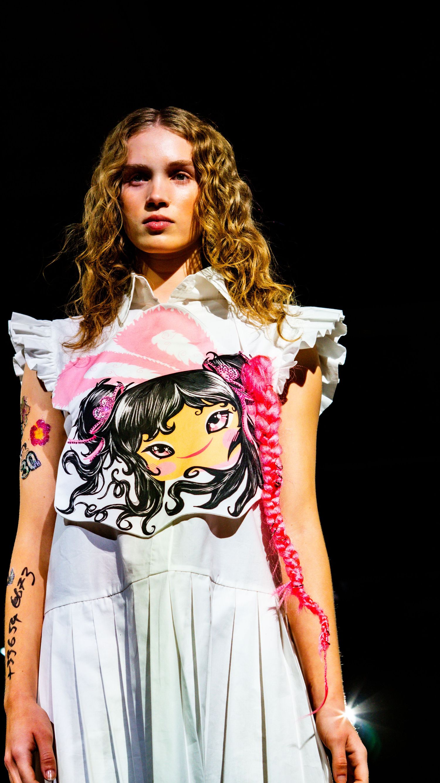 Model in white dress with anime graphic print and ruffled shoulders poses on dark fashion runway.