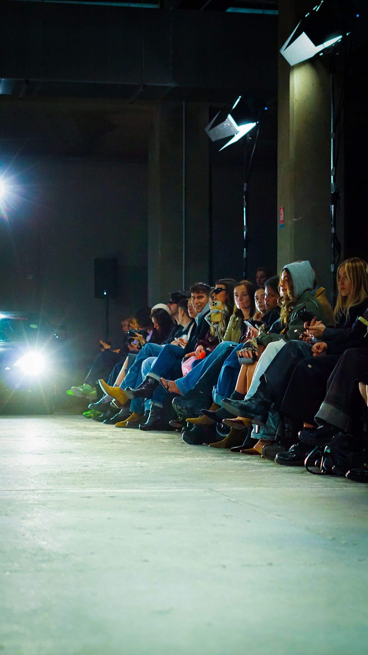 Paris fashion show audience seated along a runway lit by bright spotlights in a dark industrial venue.