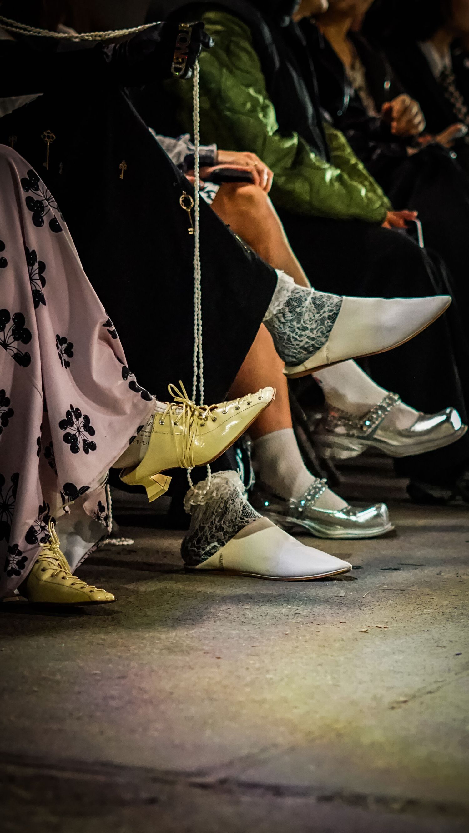 Close-up of seated fashion show attendees wearing eclectic designer shoes on a dimly lit concrete floor.