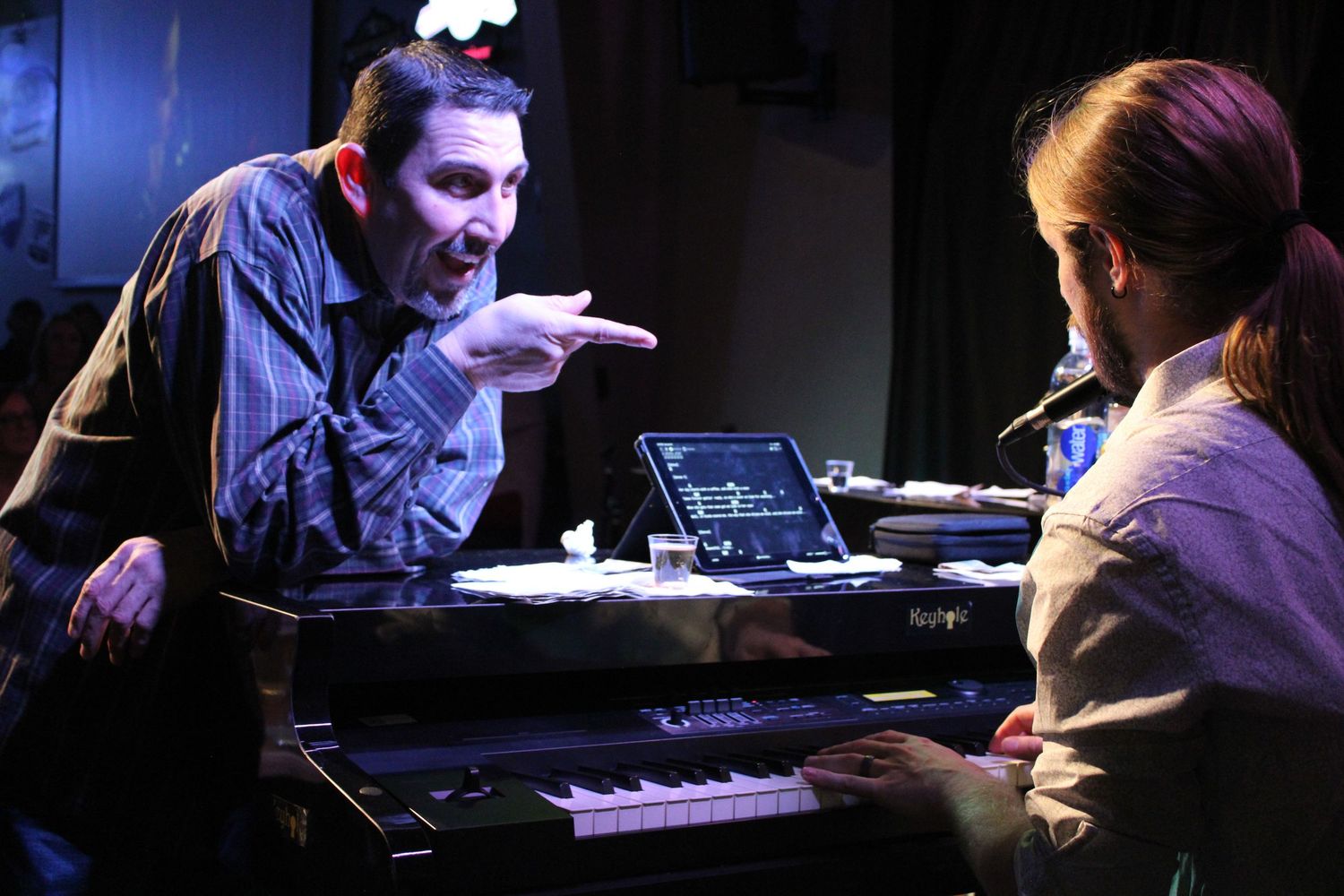 Man gesturing expressively to a man across a keyboard piano at a live show.