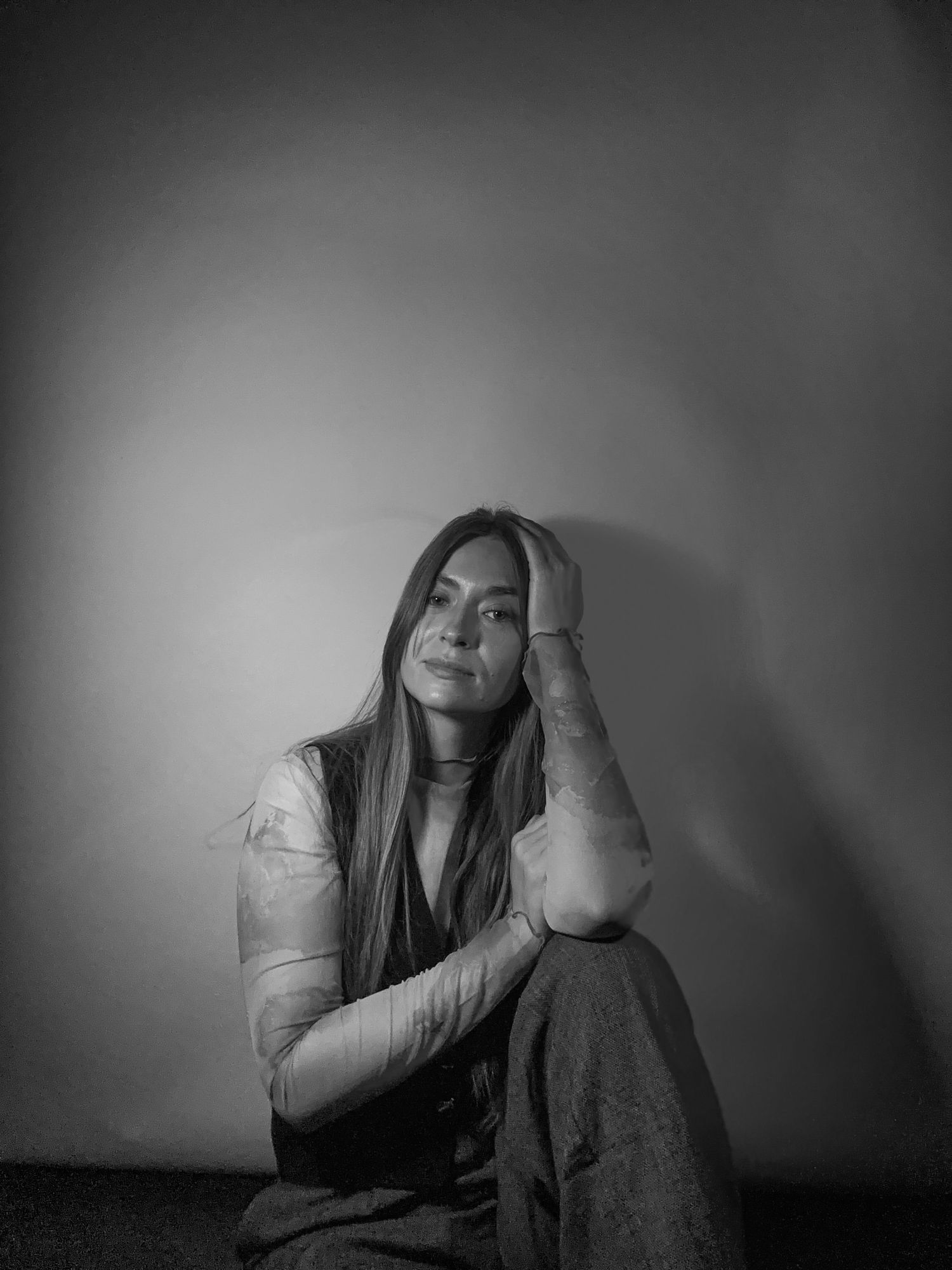 Black and white portrait of young woman sitting against wall with hand raised.