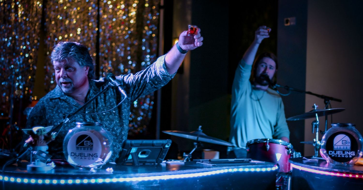 Two musicians perform on stage with colorful LED lights, one raising a drink while the other plays drums.