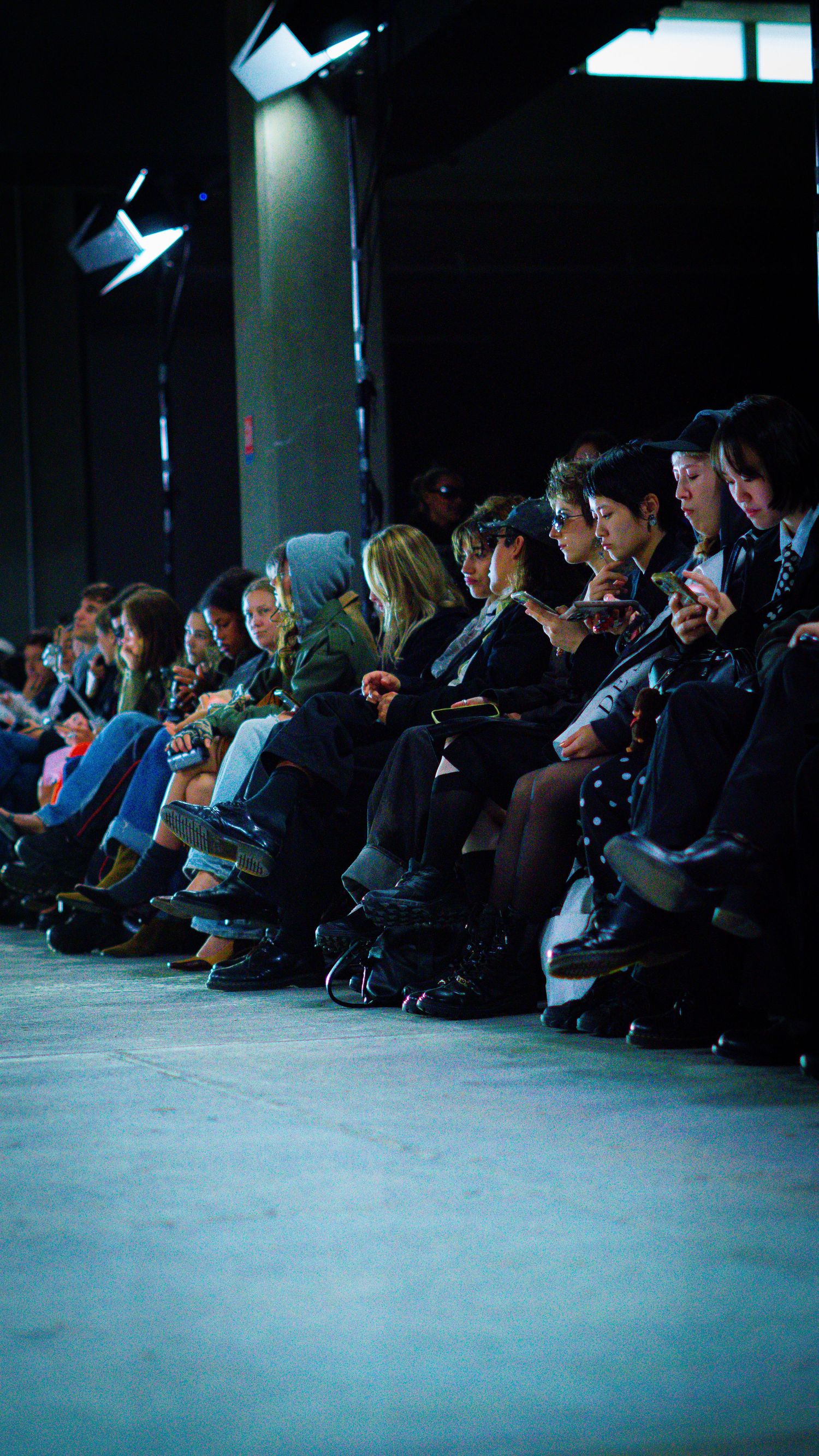 Paris fashion show front row audience seated in a dark venue, many looking at their phones.