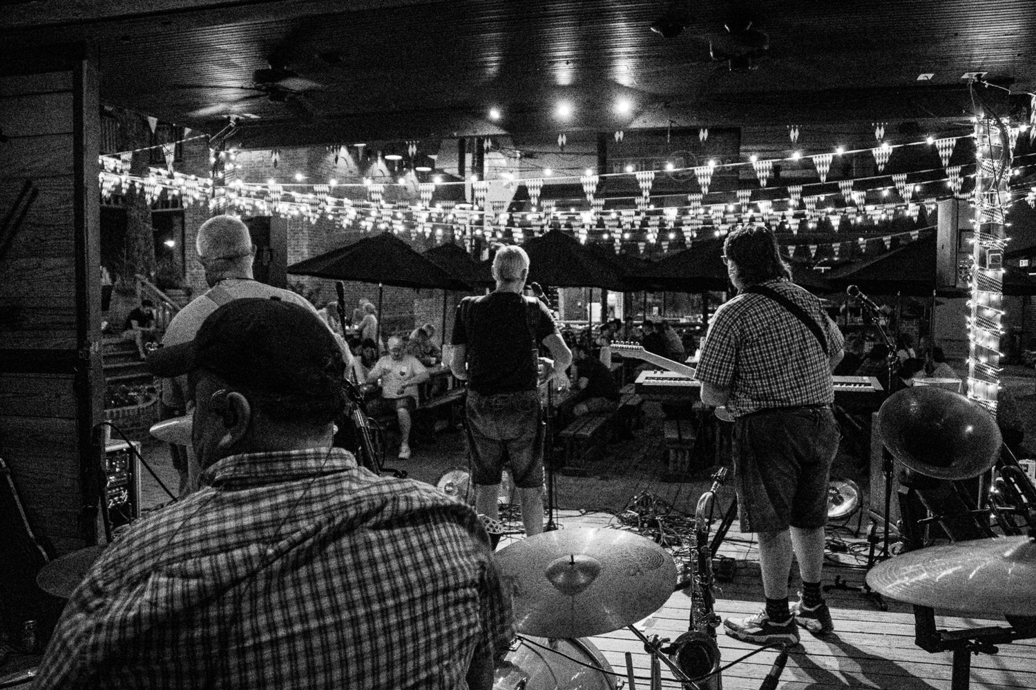 Black and white photo of a live band performing on an outdoor stage with string lights overhead.