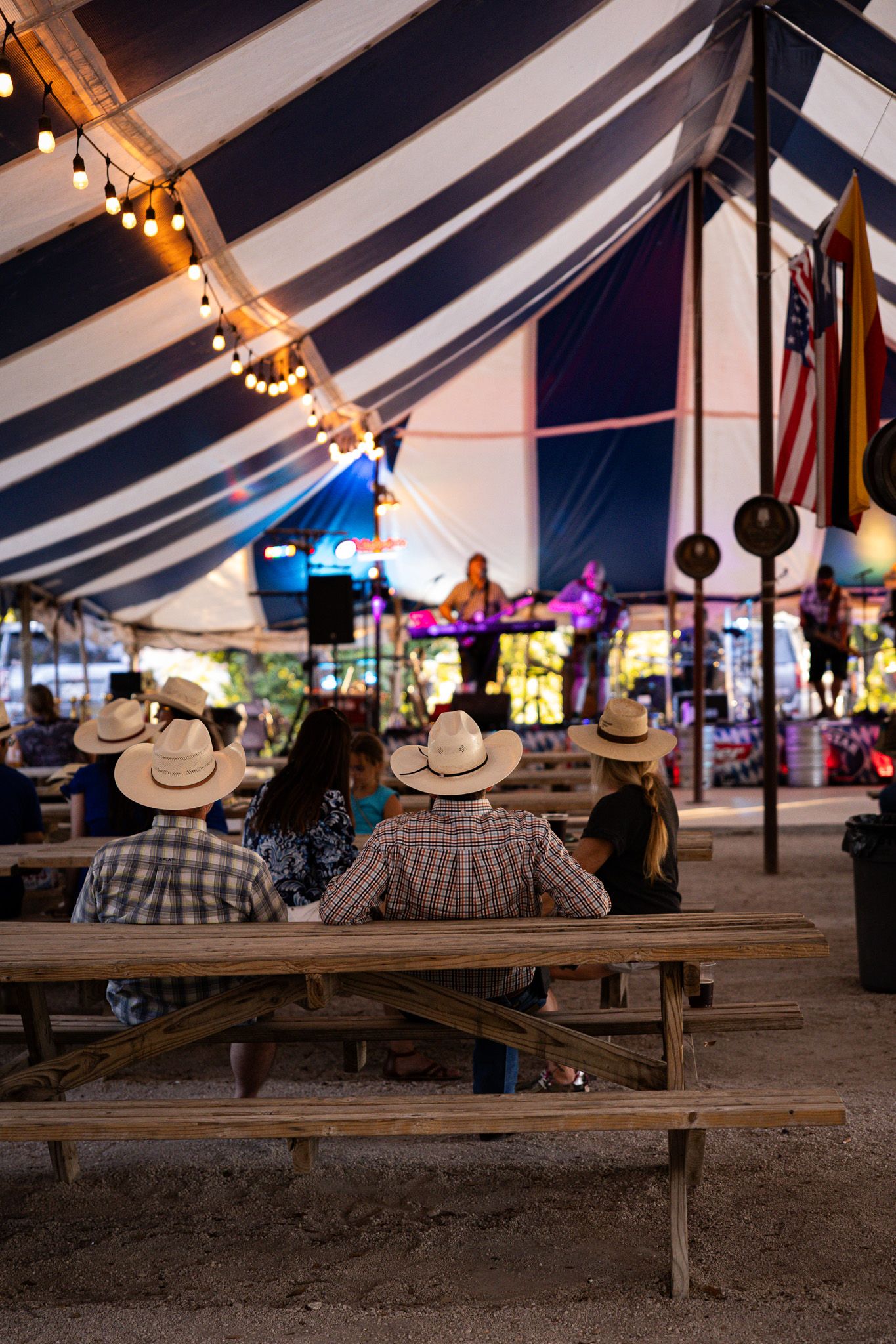 People wearing cowboy hats seated at wooden picnic tables inside a blue and white striped tent at a German festival.