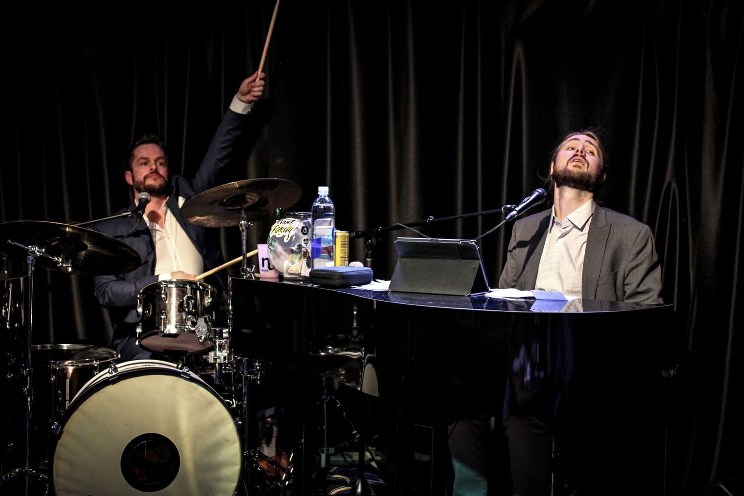 Two male musicians performing live, one playing drums and one singing at a piano on a dark stage.