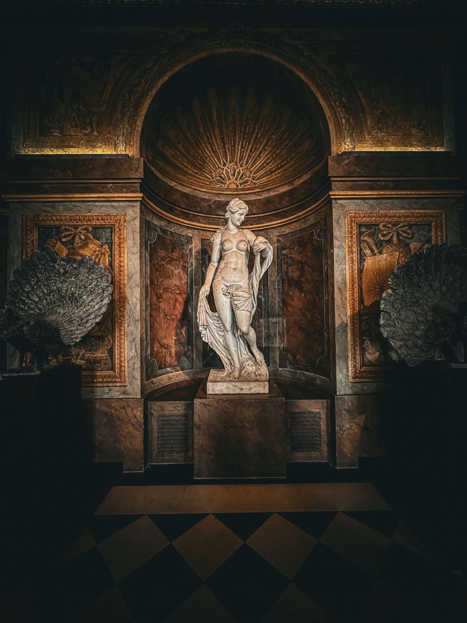 Classical marble statue displayed in ornate golden niche flanked by peacock feathers.