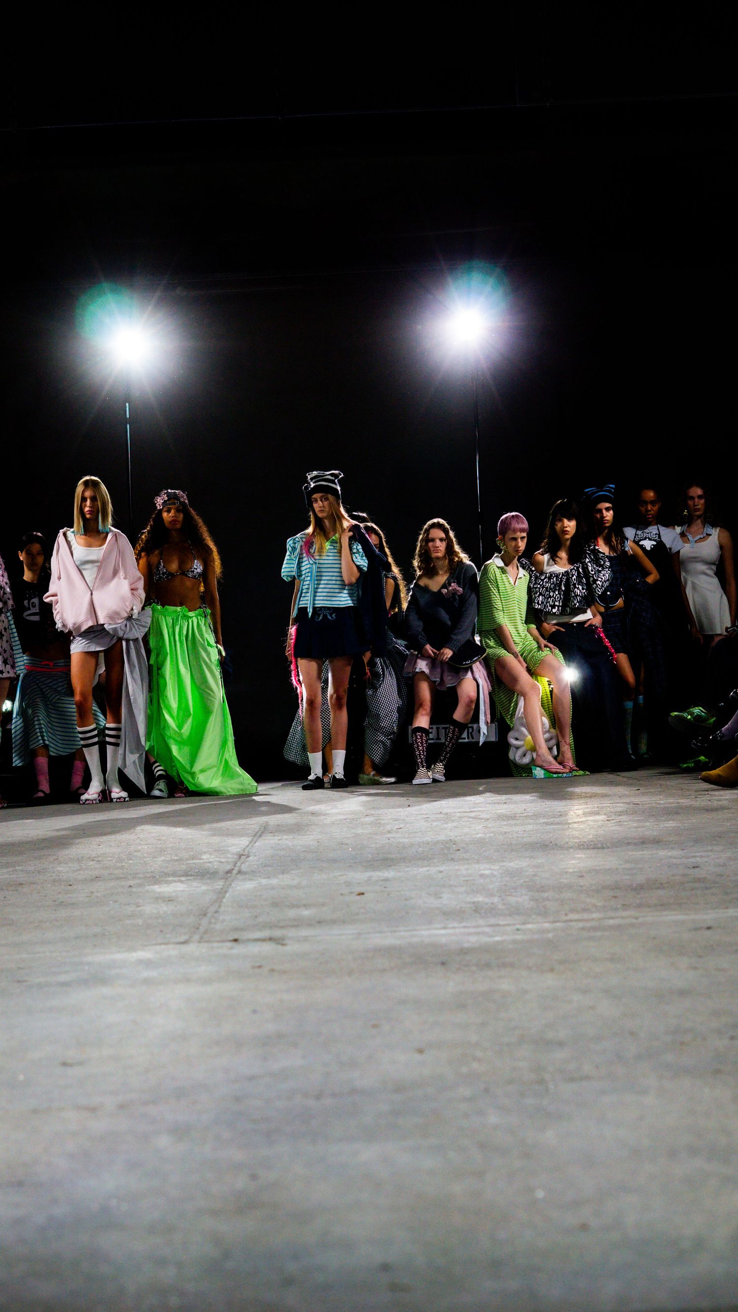 Models walk the runway at a Paris night fashion show under bright spotlights wearing colorful avant-garde outfits.