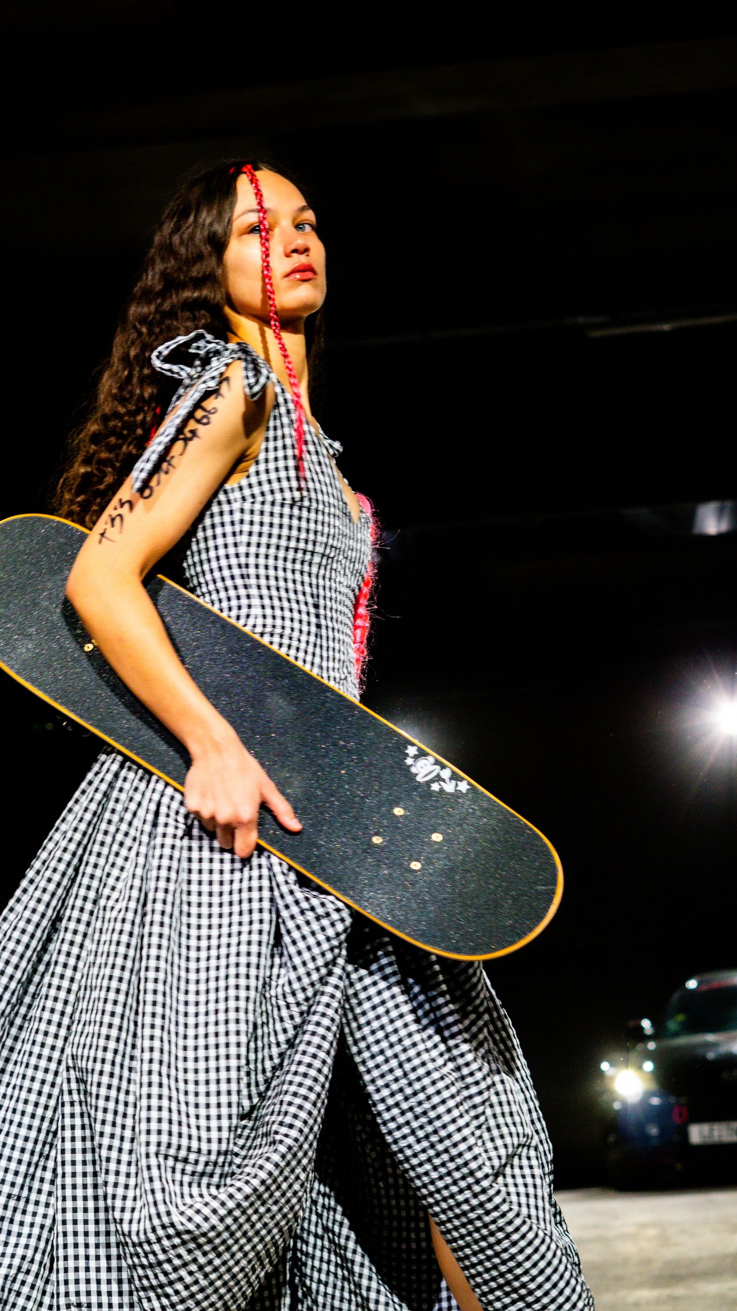 Model in gingham dress carries a skateboard on a dramatic dark runway with car headlights behind.