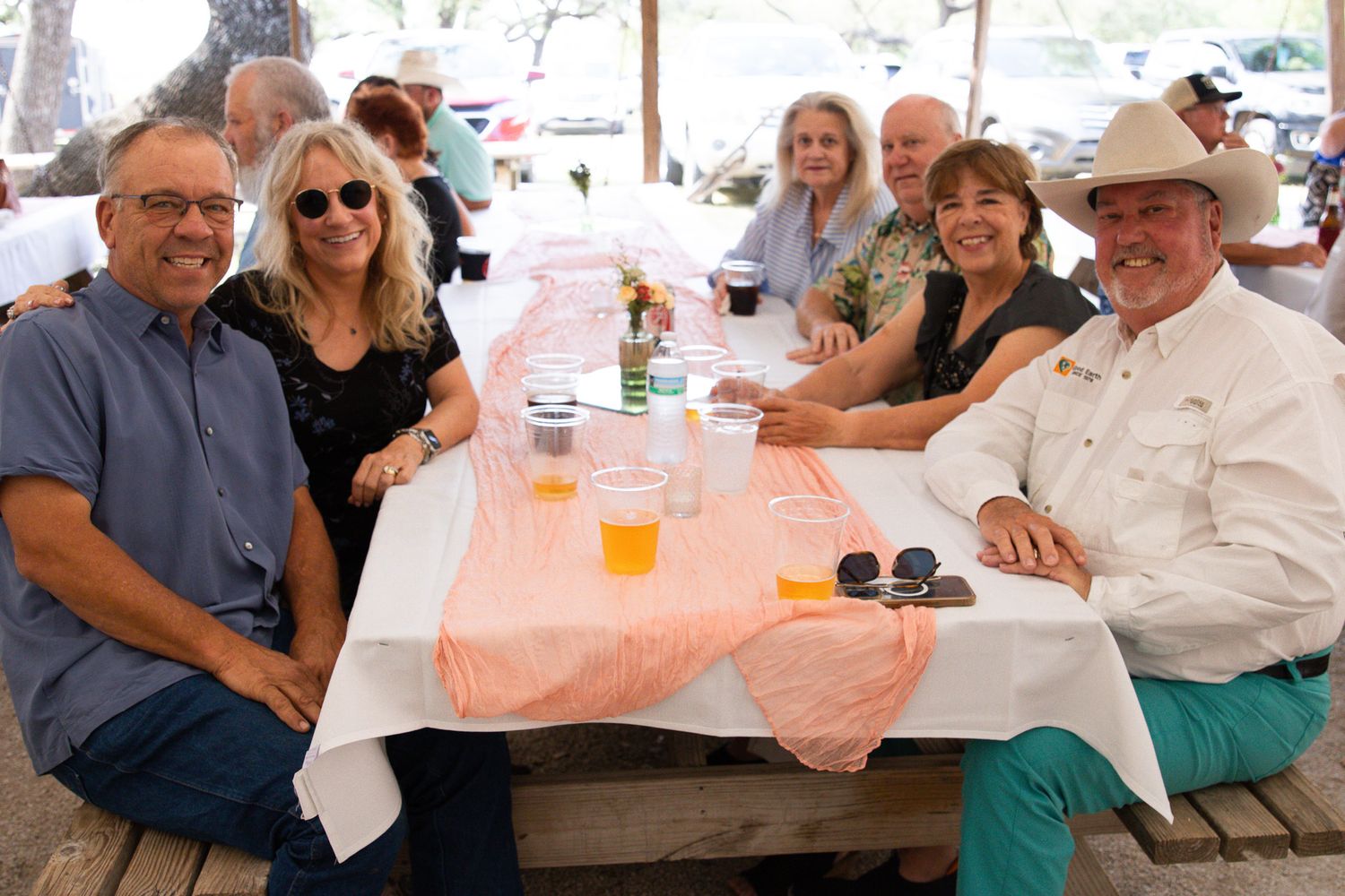 Group of five adults smiling and sitting together at a picnic table at an outdoor event.