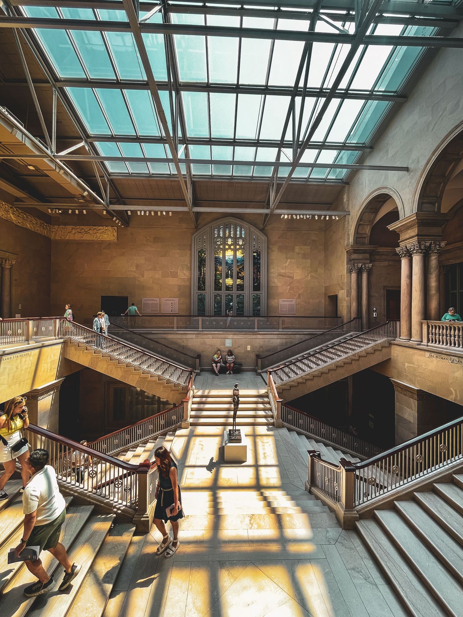 Sunlit grand museum interior with glass ceiling, staircases, and visitors exploring.
