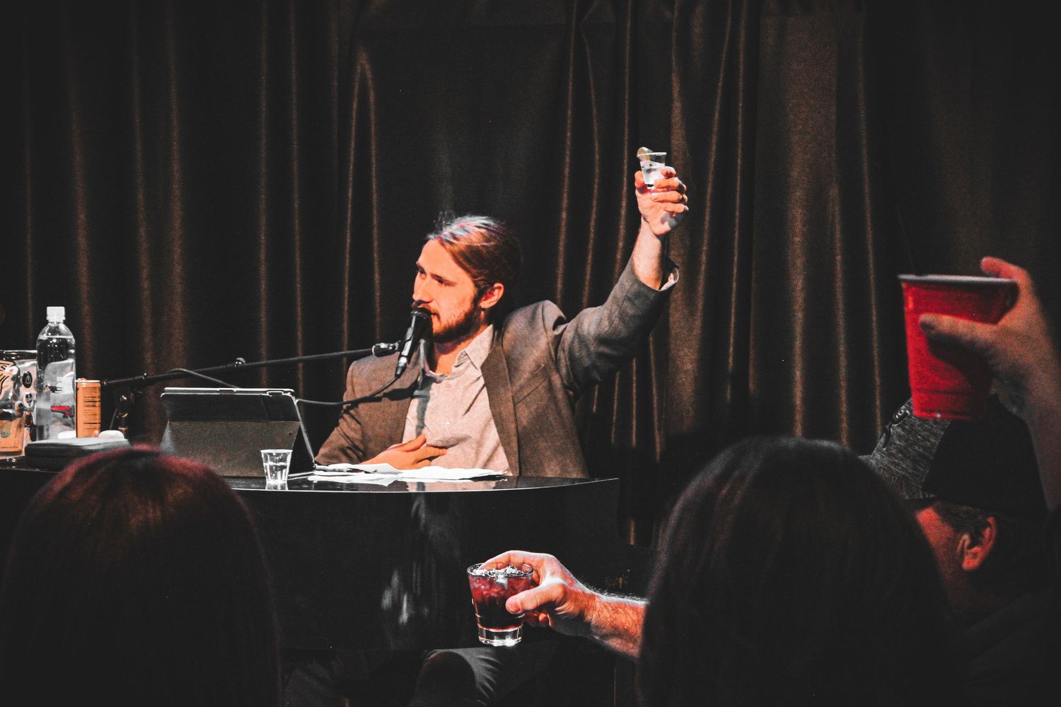 Bearded man in suit raising a glass at a dimly lit piano bar event.
