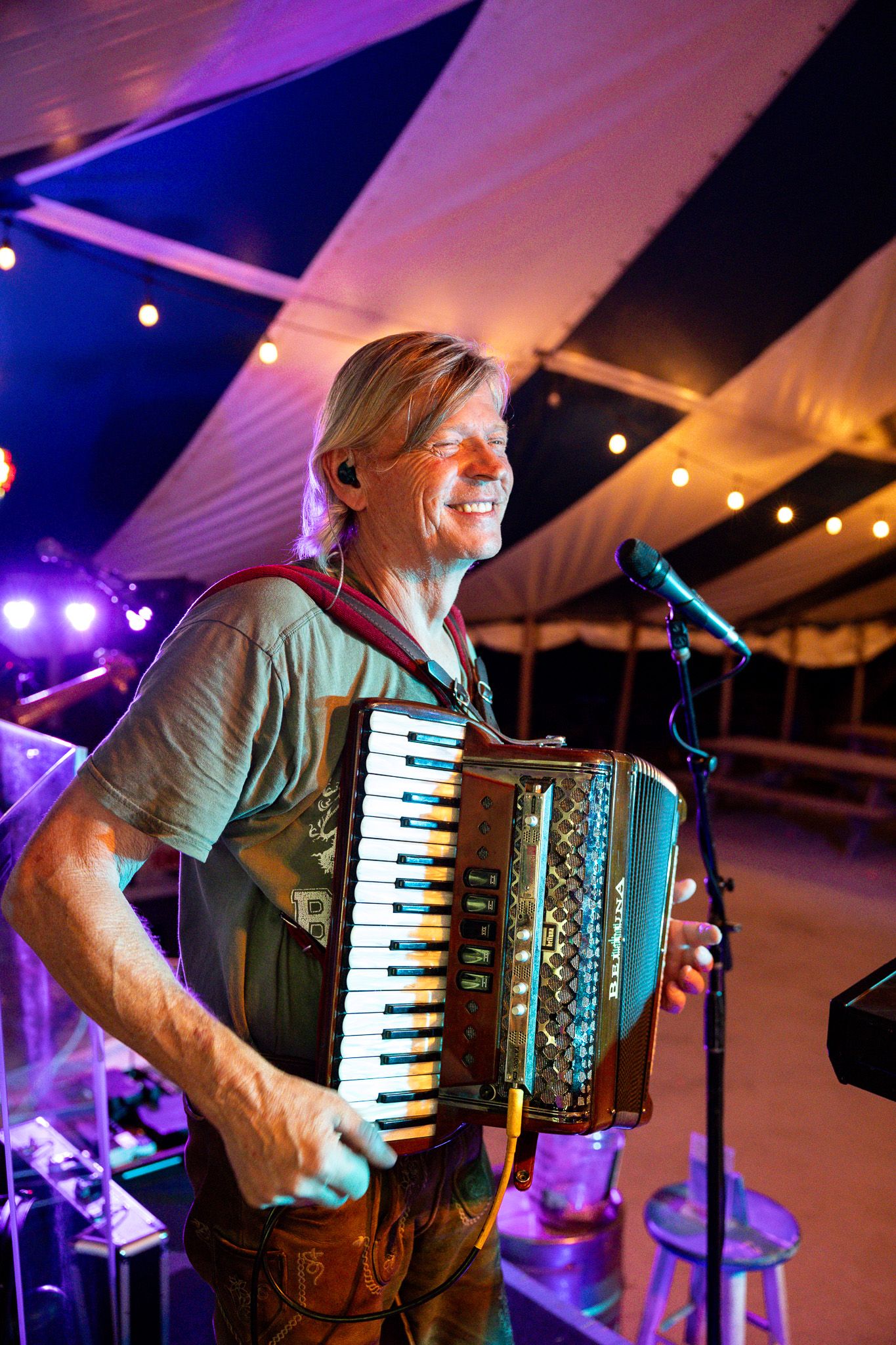 Smiling blonde musician playing an accordion on stage under colorful lights inside a festival tent.