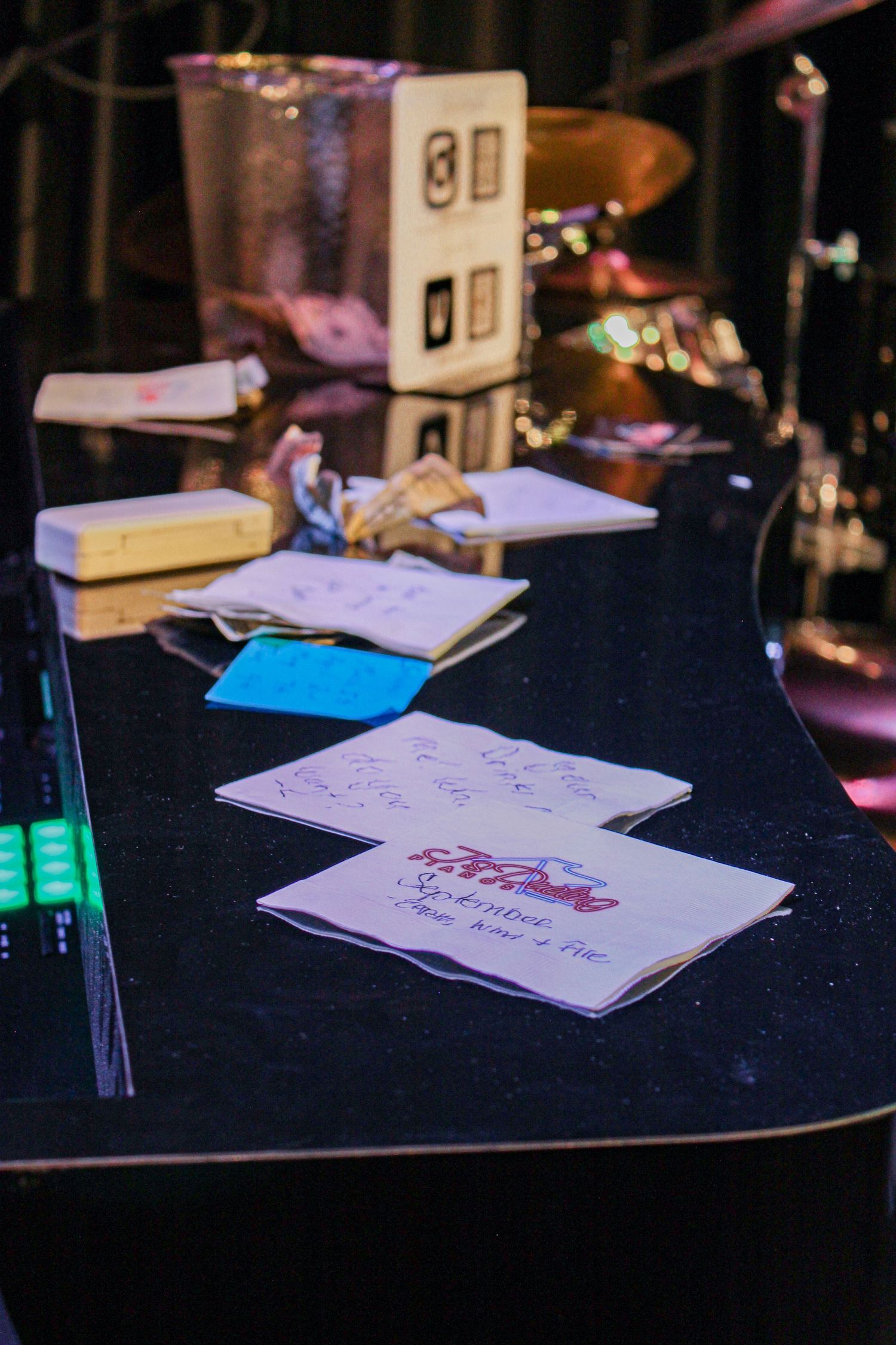 Music stage setup with handwritten notes and setlists on a black piano with drums in background.