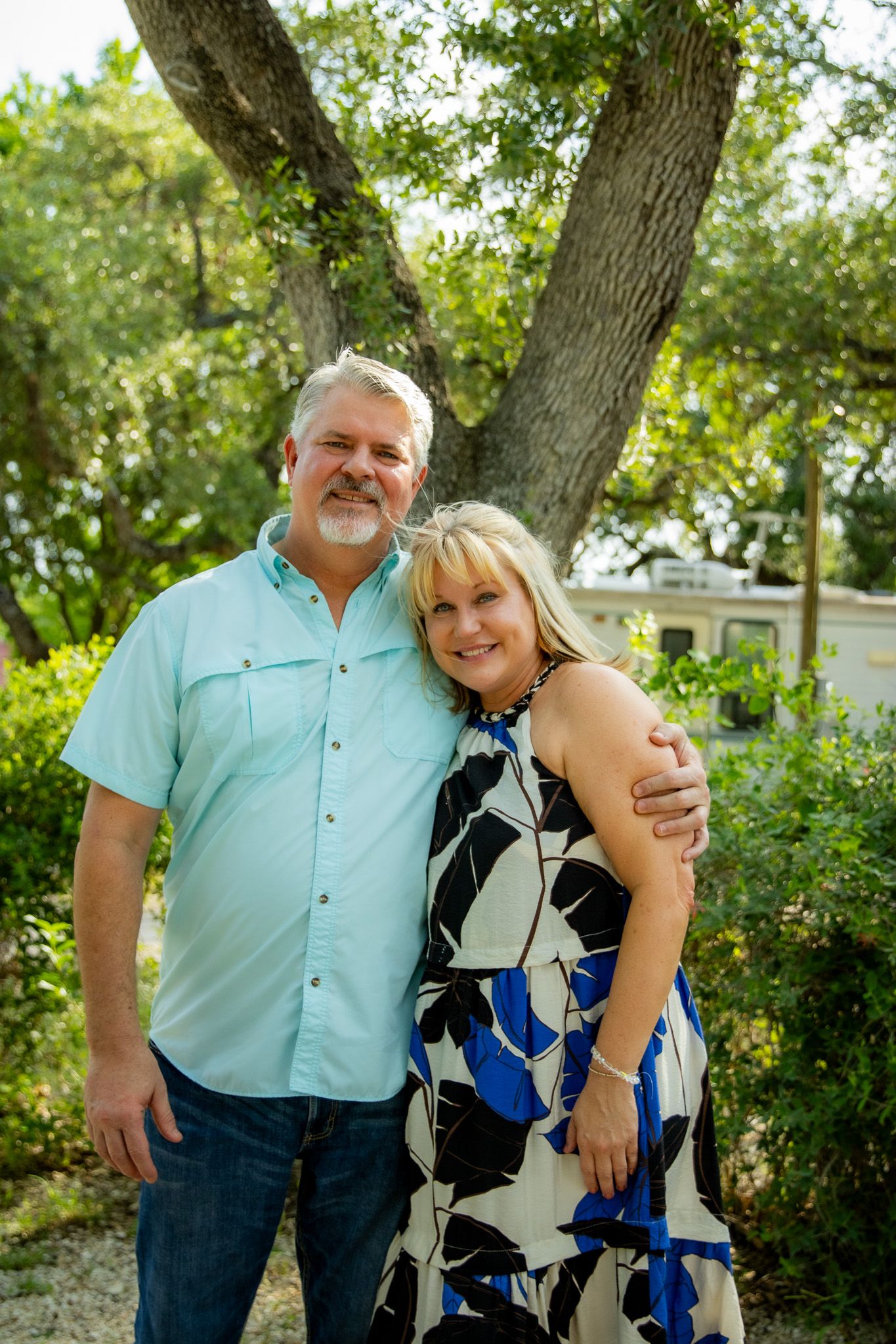 Smiling couple poses outdoors under a large oak tree surrounded by lush greenery.