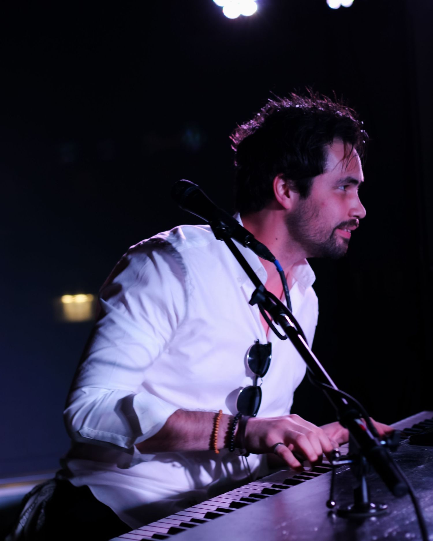 Male musician plays keyboard on stage in a white shirt under dramatic stage lighting.