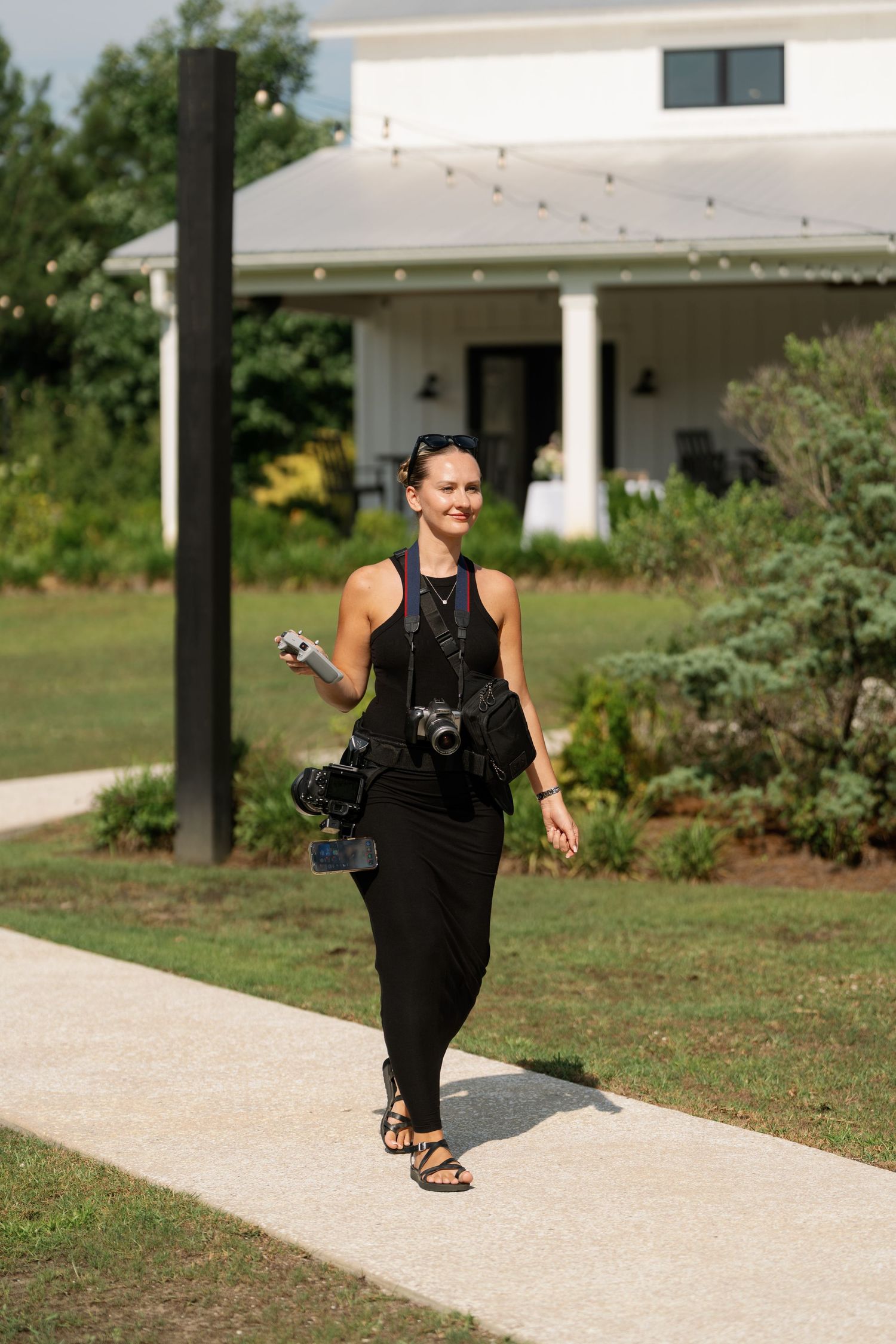A smiling female photographer in all-black outfit walks on a path carrying cameras, with a white farmhouse in the background.
