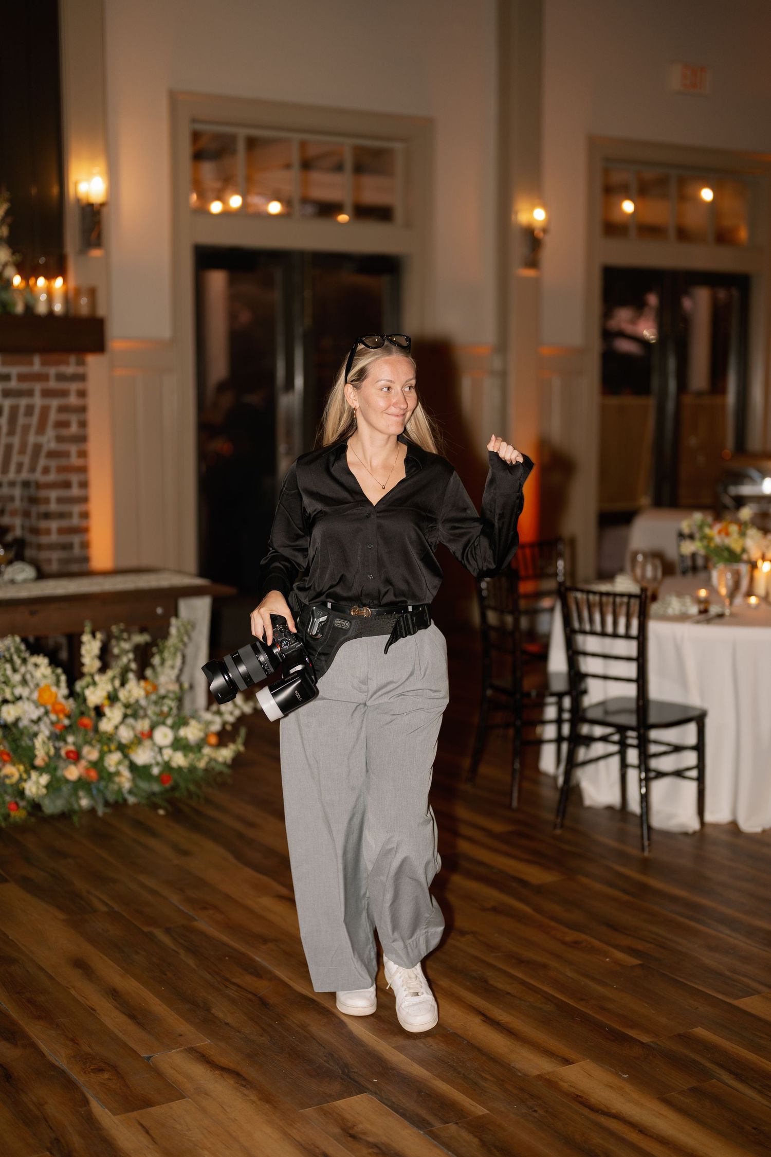 A female photographer walks through an elegant wedding reception venue holding a camera, wearing a black blouse and gray trousers.
