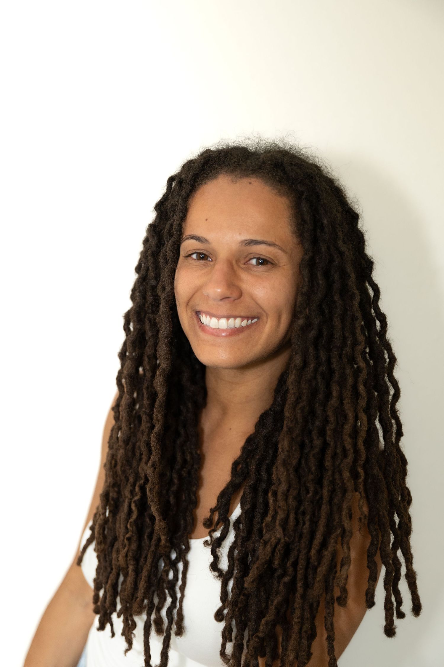 Smiling woman with long brown dreadlocks wearing a white top against a white background.