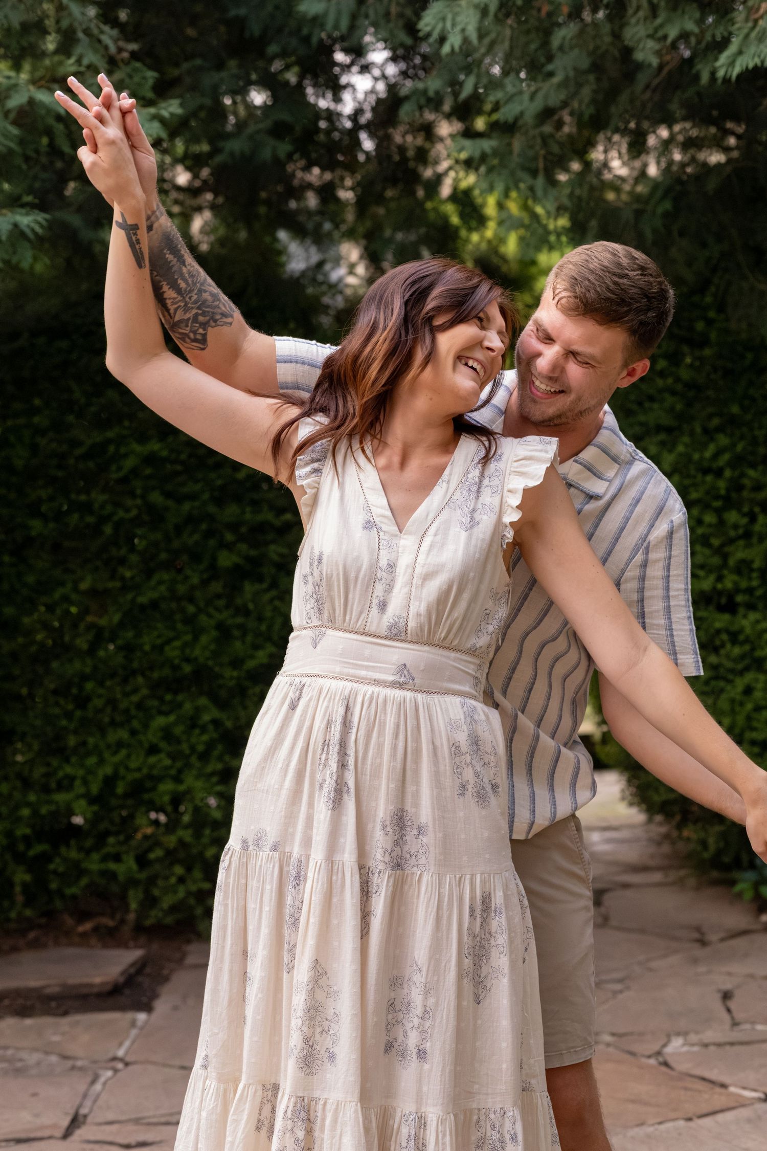 A joyful couple dancing outdoors, woman in floral dress with tattoos laughing, man in striped shirt, lush green garden backdrop.