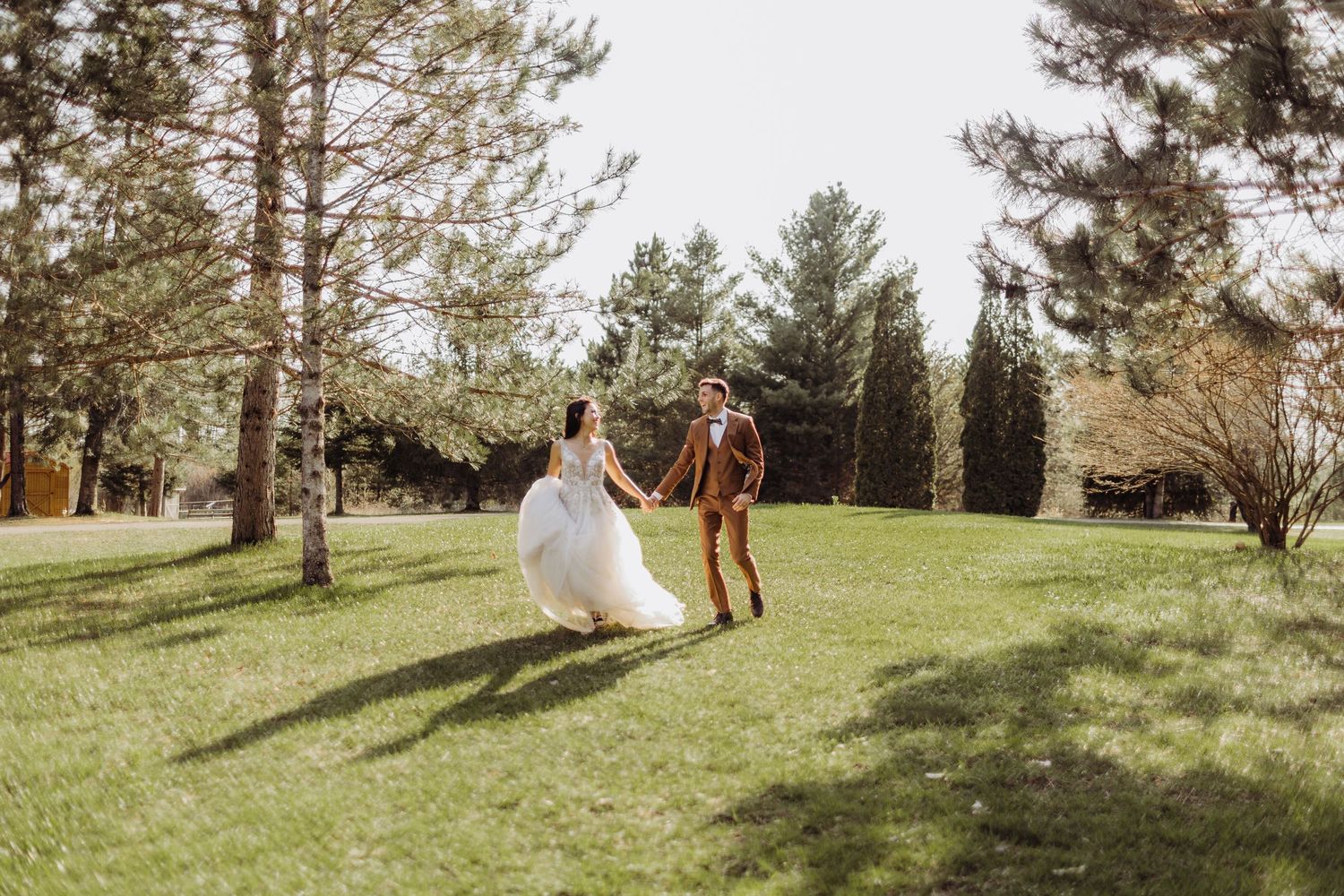 a bride and groom hold hands while running though the grass between pine trees in the sun.