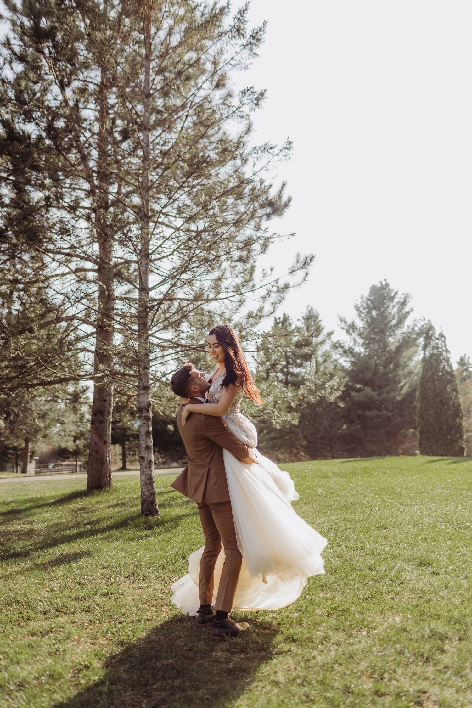 a bride and groom, the groom lifts the bride and spins her in the grass between pine trees in the sun.
