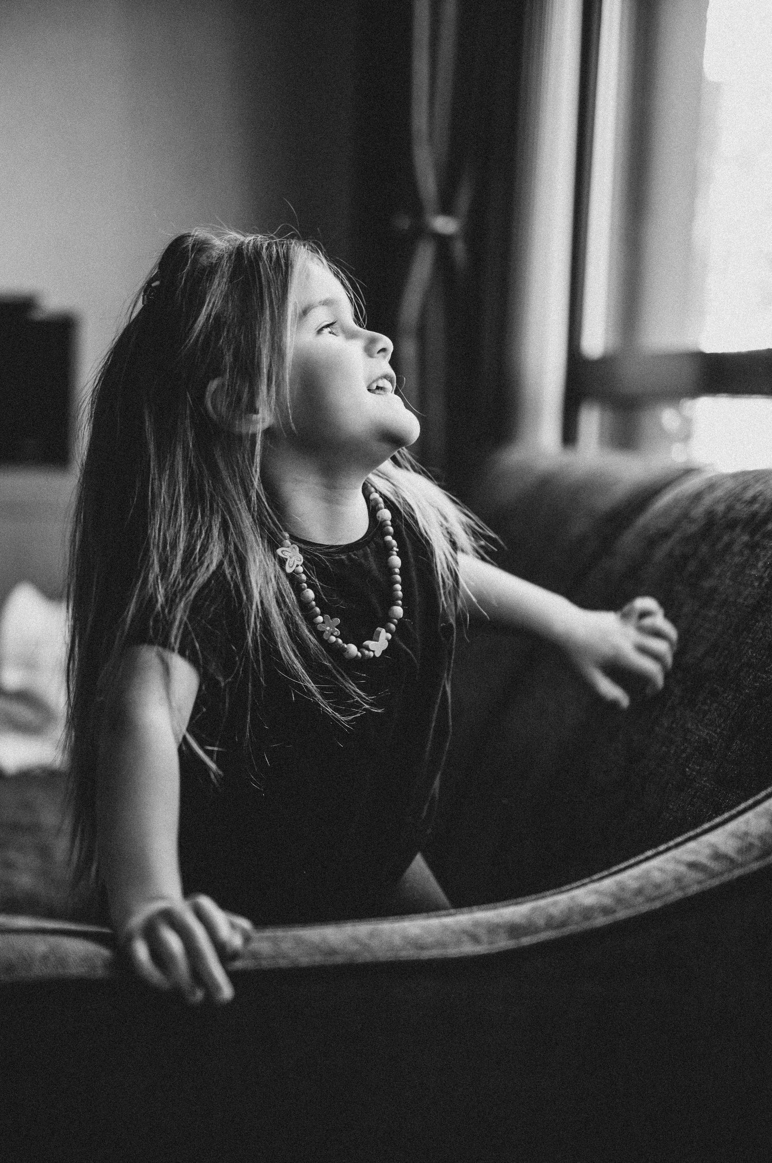 A black and white  portrait of a girl looking out of the window, documentary-style family photography capturing everyday life