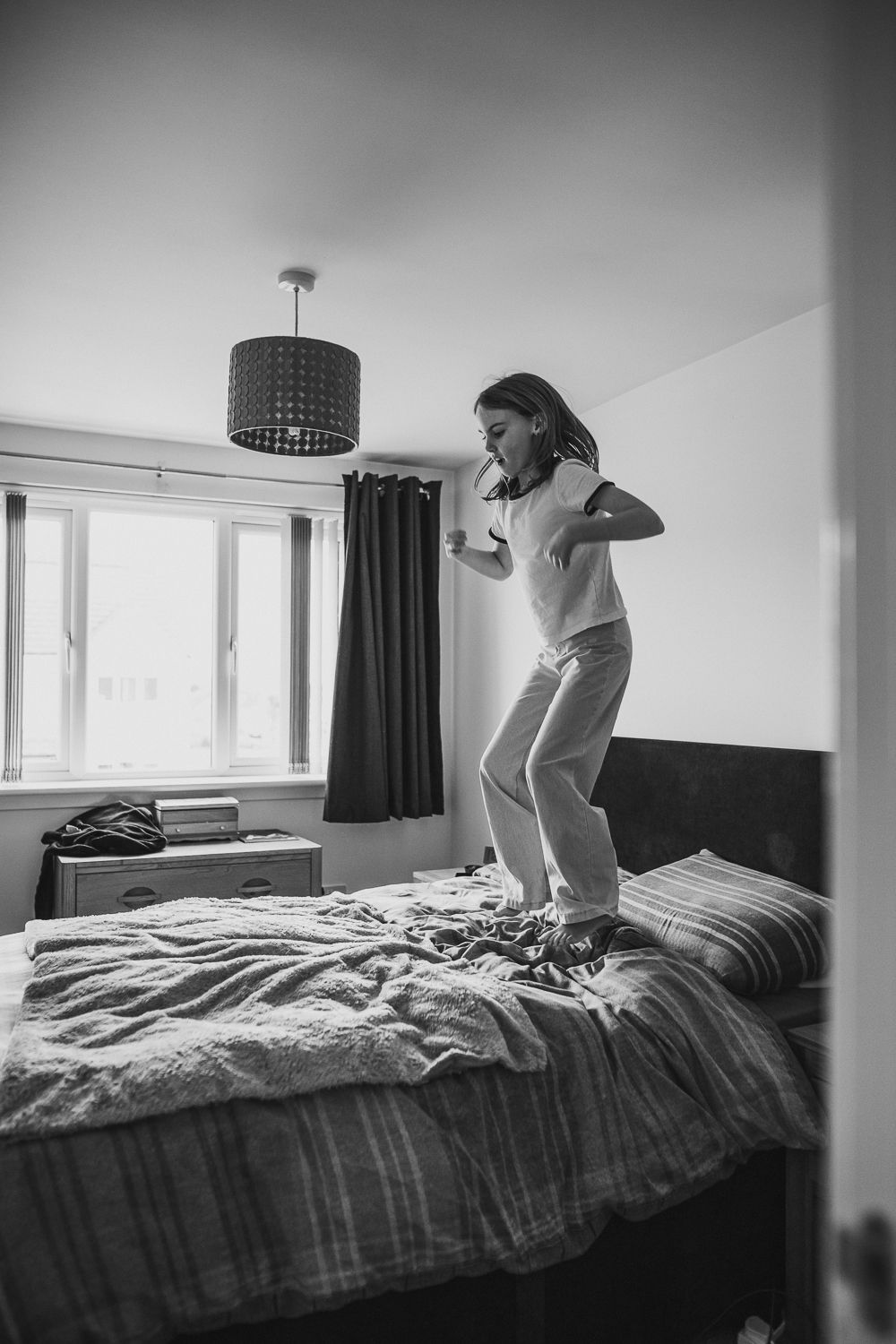 Black-and-white photo of child jumping on a bed indoors, capturing playful everyday moments.