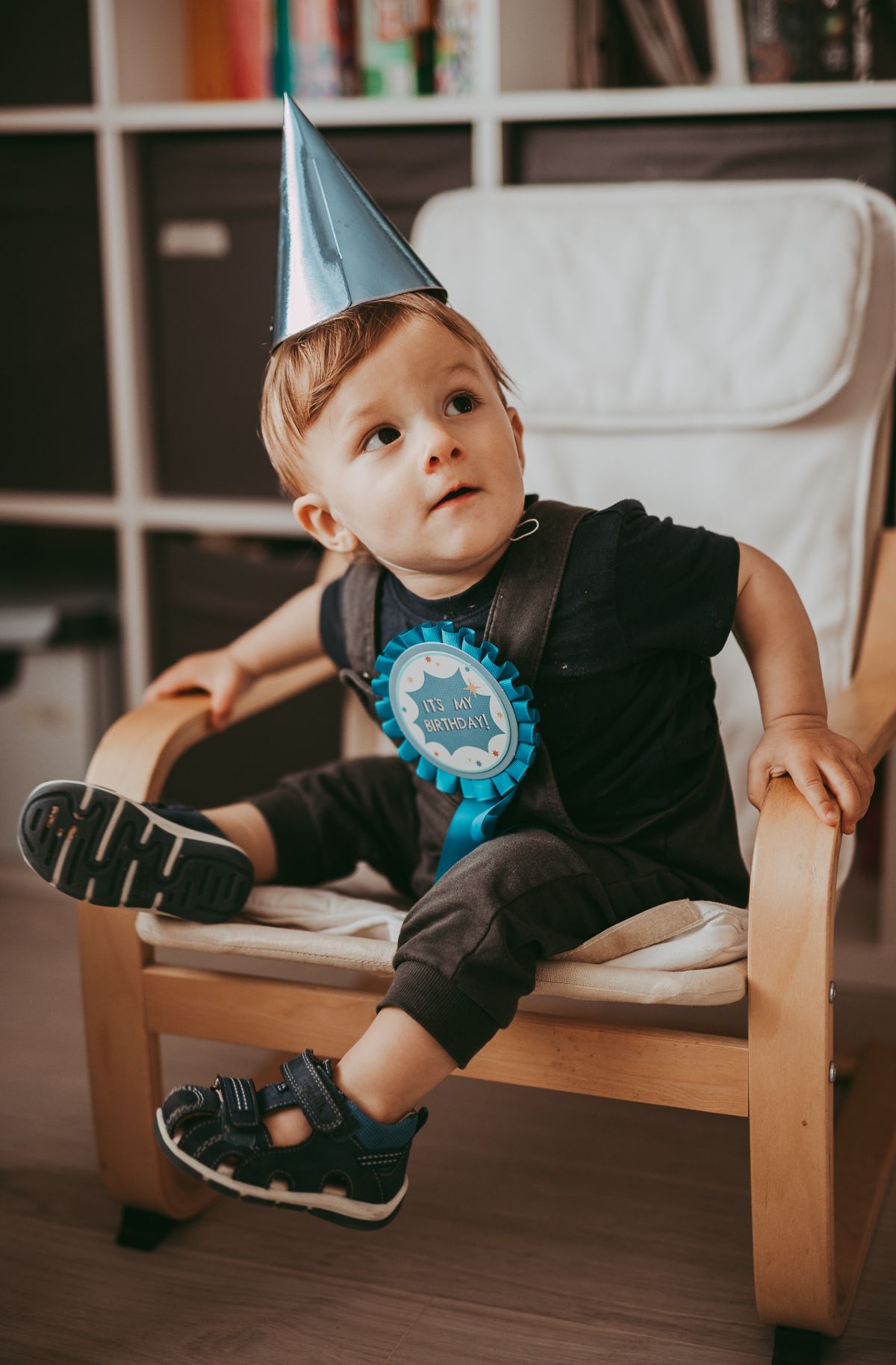 A boy wearing a birthday hat and a  badge captured during a relaxed birthday family photoshoot with toddler and parents indoors