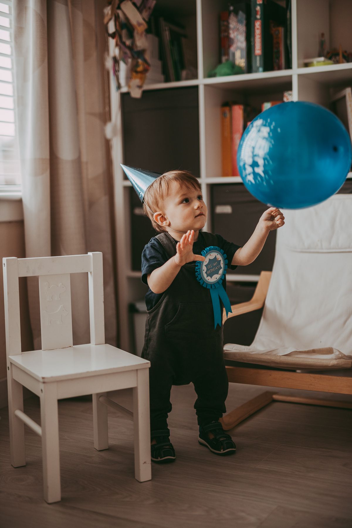 A toddler with a birthday hat, badge and a balloon celebrating 1st birthday captured during a family photoshoot indoors