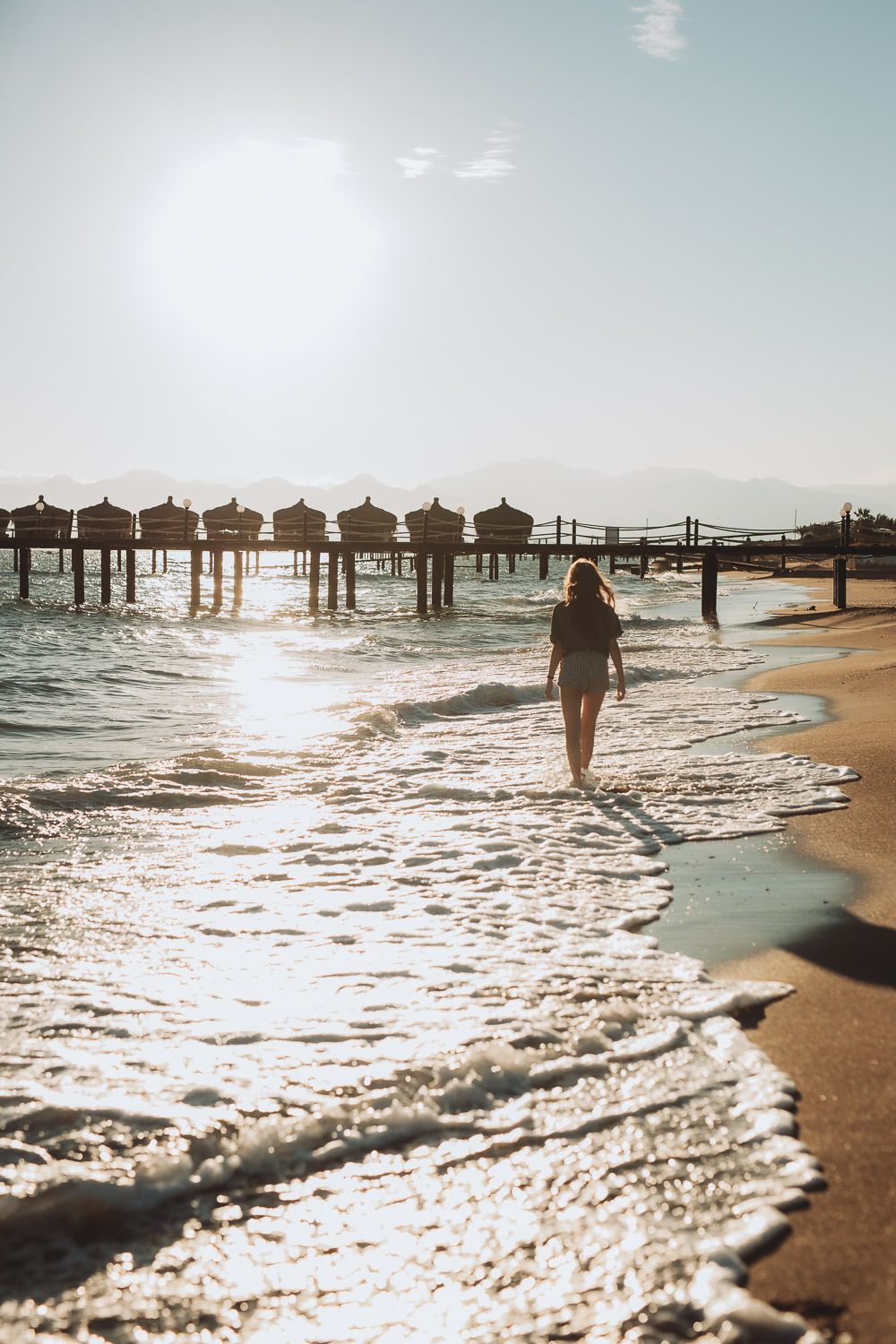 Child walking along the shoreline with waves and pier in the background, relaxed beach photography.