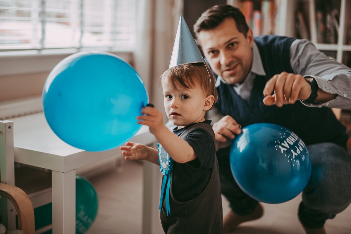 A toddler with a birthday hat, badge and a balloon with a parent celebrating 1st birthday captured during a family photoshoot indoors