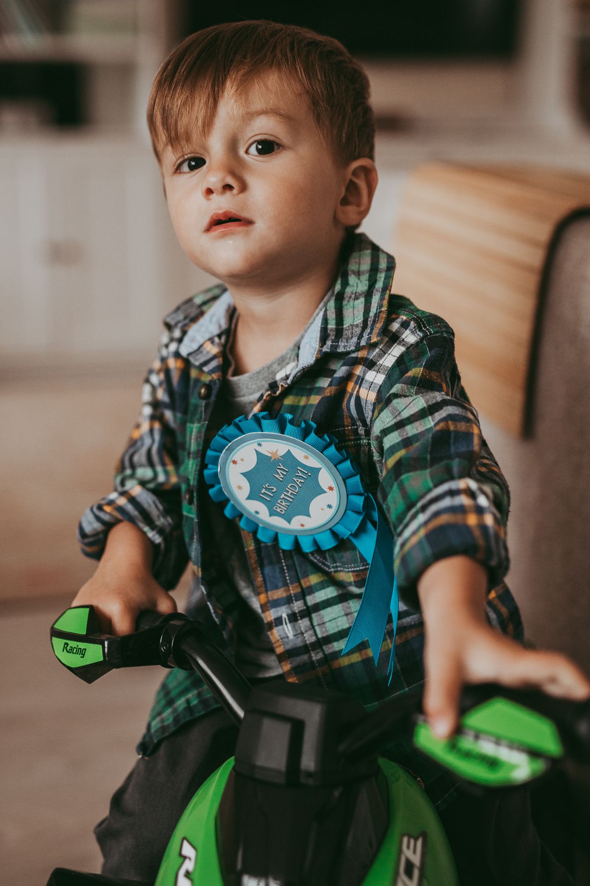 A boy riding a motorbike indoors and wearing a birthday badge, relaxed birthday family photoshoot with toddler and parents indoors