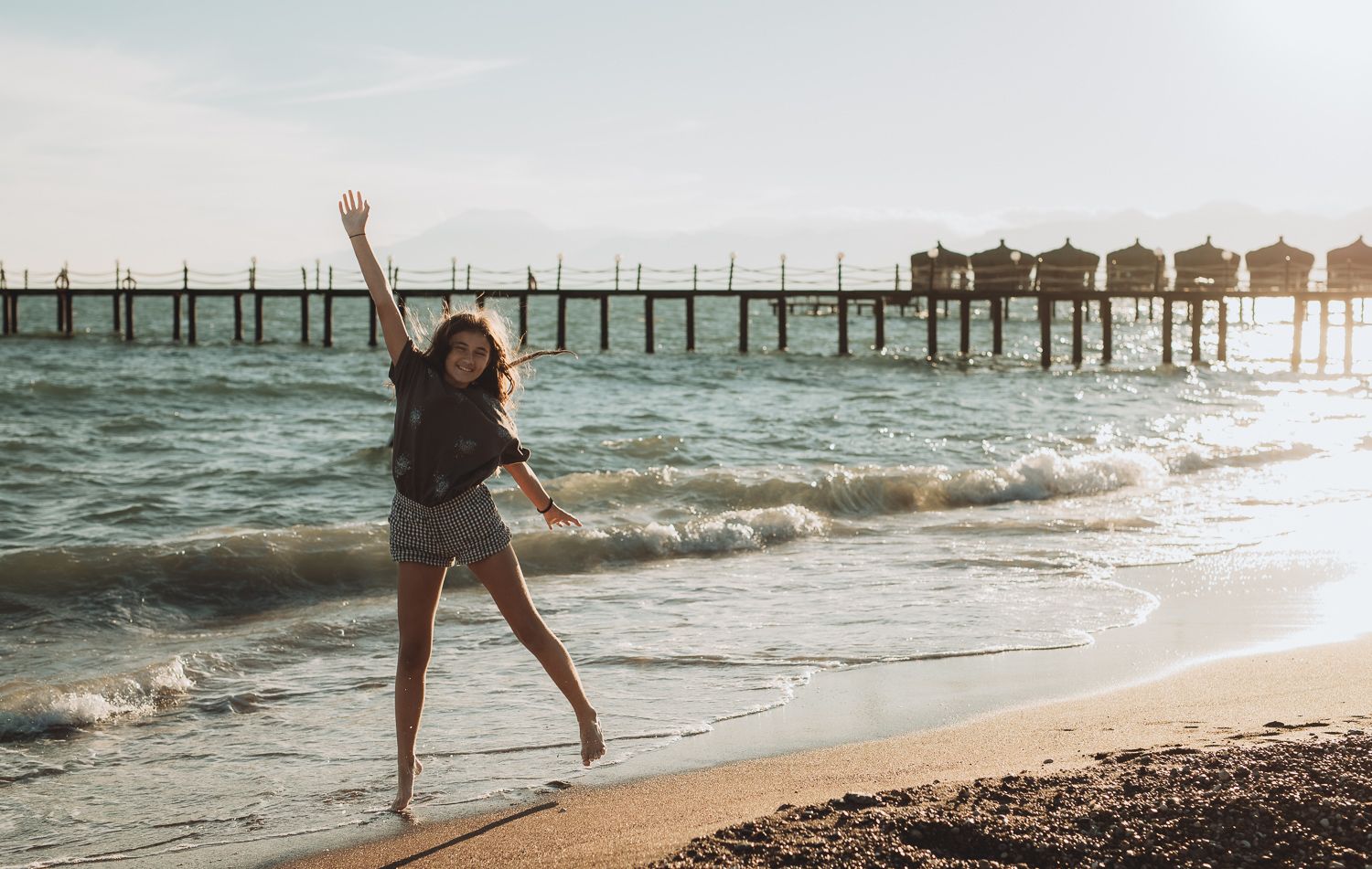 Child jumping joyfully in the shallow sea with a pier in the background, lifestyle family photography