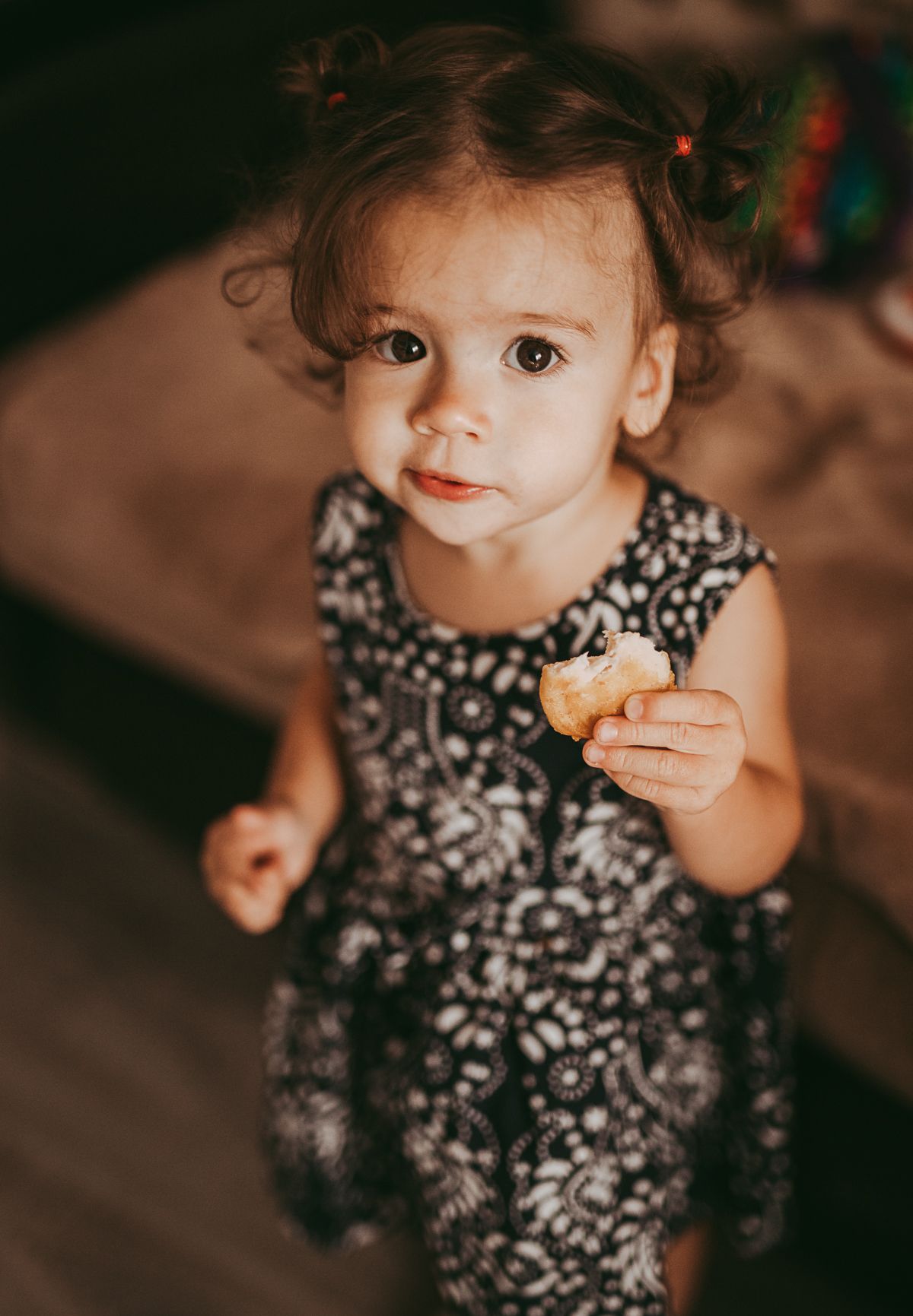 Toddler girl holding a cookie and looking at the camera indoors, lifestyle portrait capturing everyday moments.