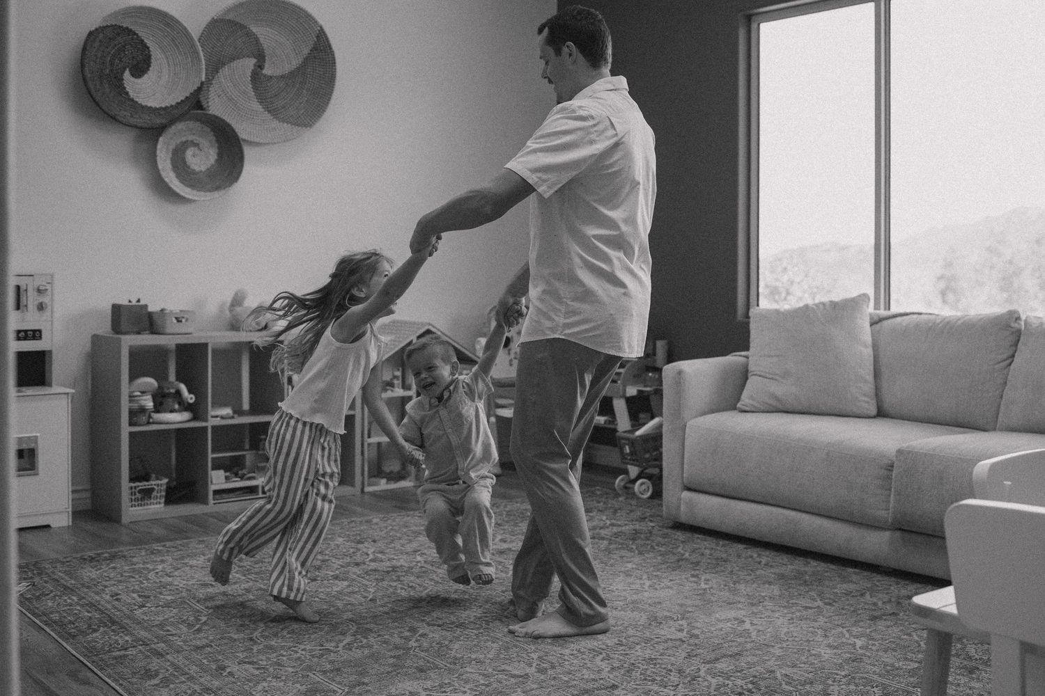 Black and white photo of a father dancing with two young children in a cozy living room.
