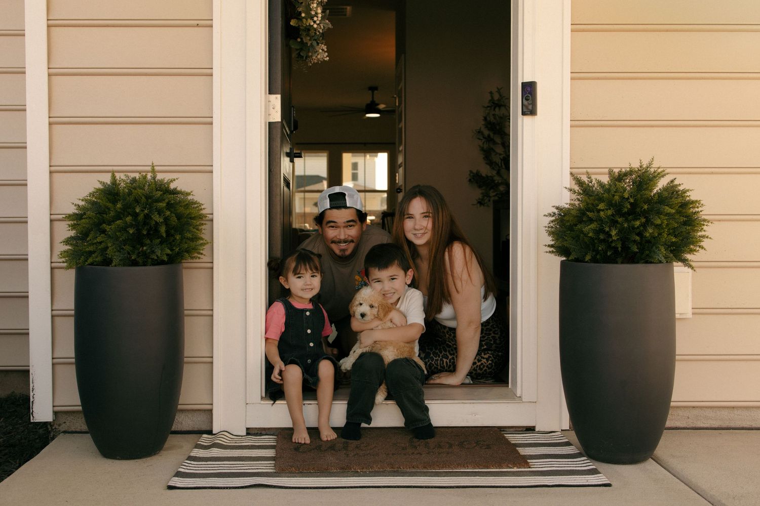 Family of four with a puppy posing happily at their front door flanked by tall dark planters with green shrubs.