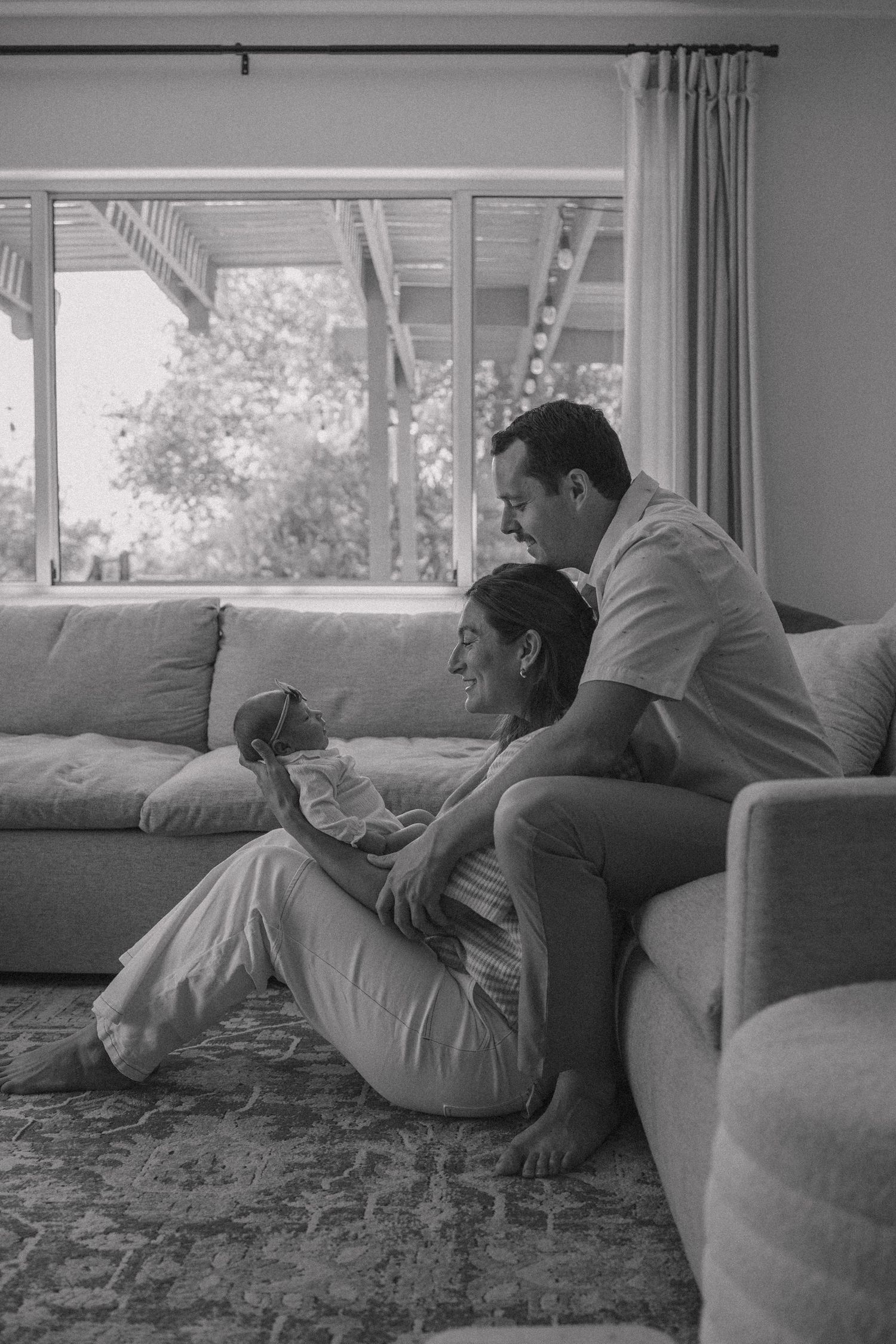 Black and white photo of couple sitting on living room floor leaning against sofa admiring their newborn baby.