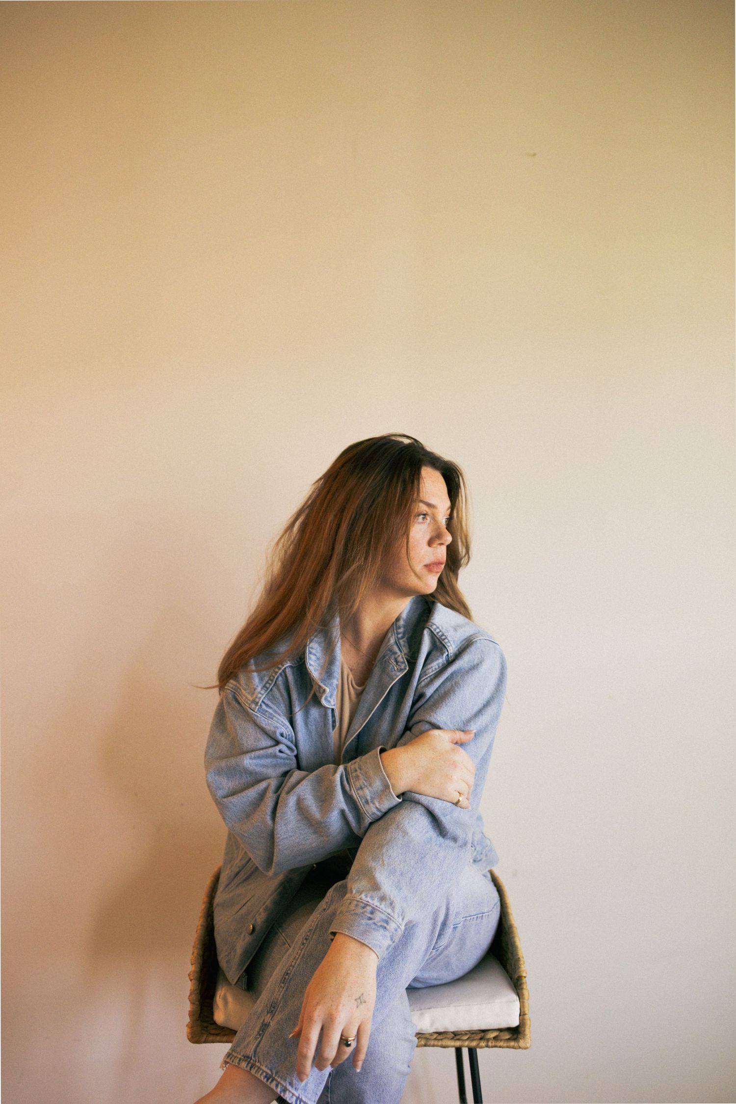 A person in a light denim jacket poses artistically against a beige wall in a series of moody editorial shots.