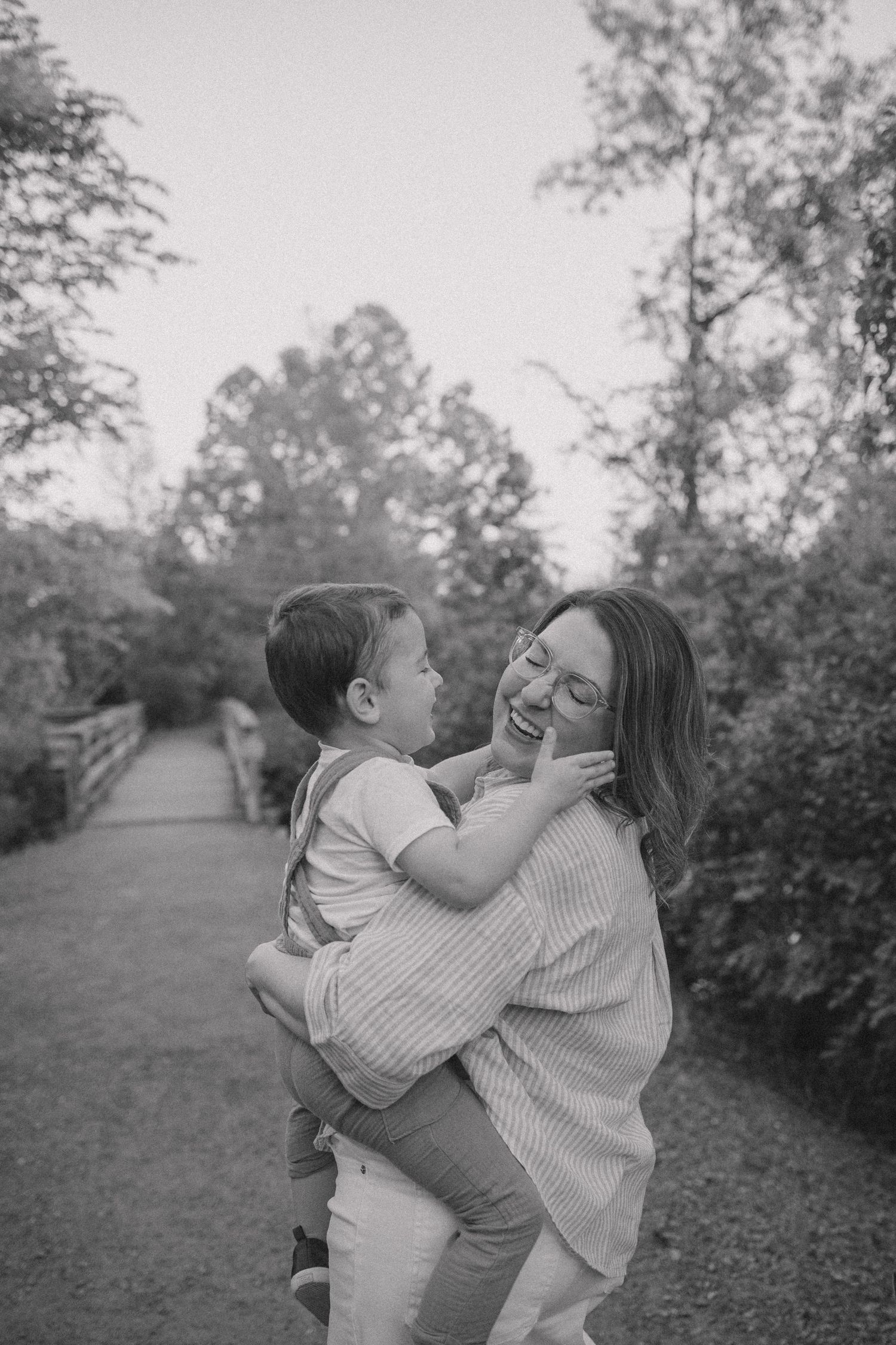 Black and white photo of a laughing mother holding her young son outdoors on a garden path surrounded by trees.
