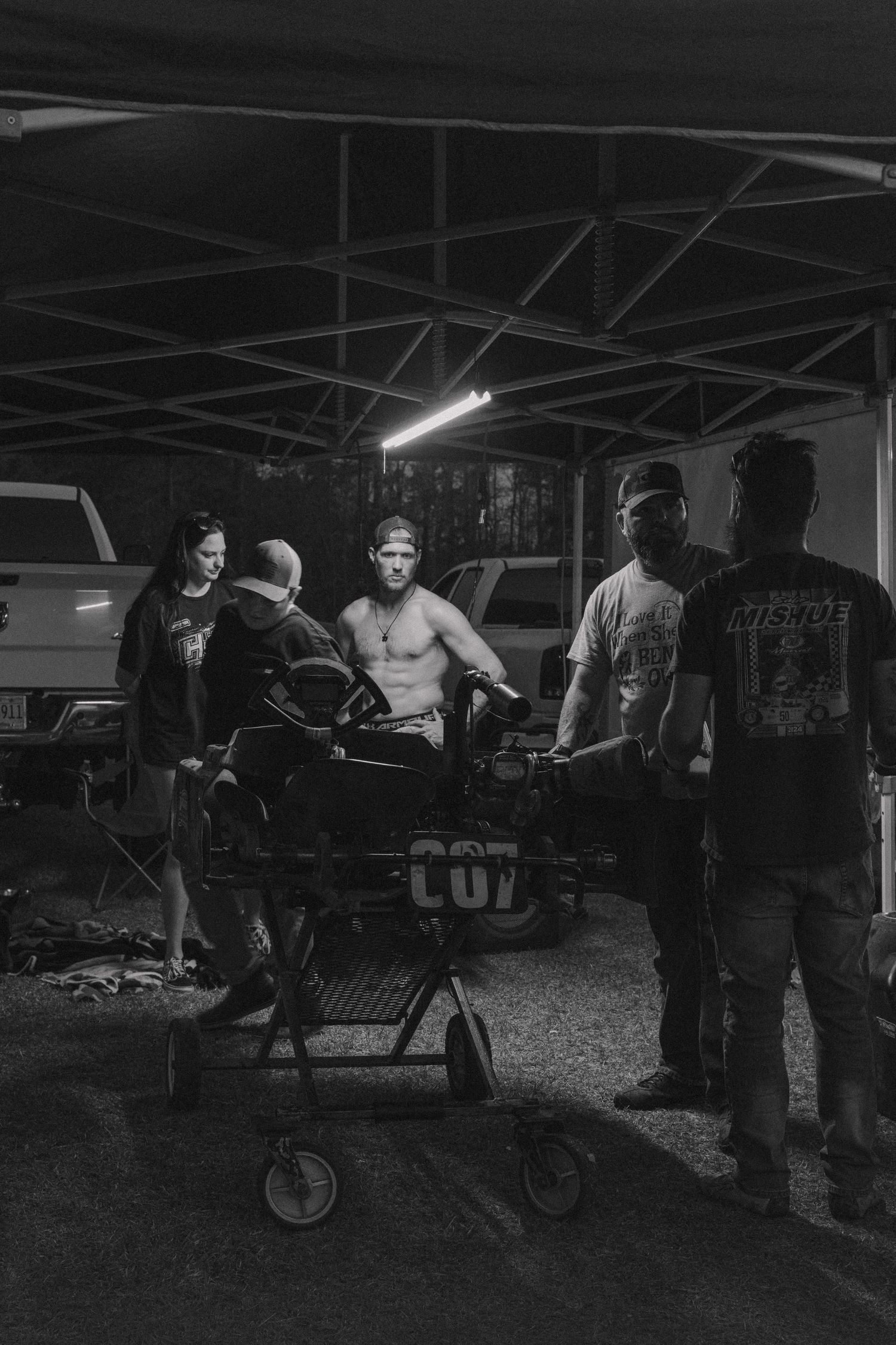 Black and white photo of shirtless man on stretcher surrounded by people under canopy at night.