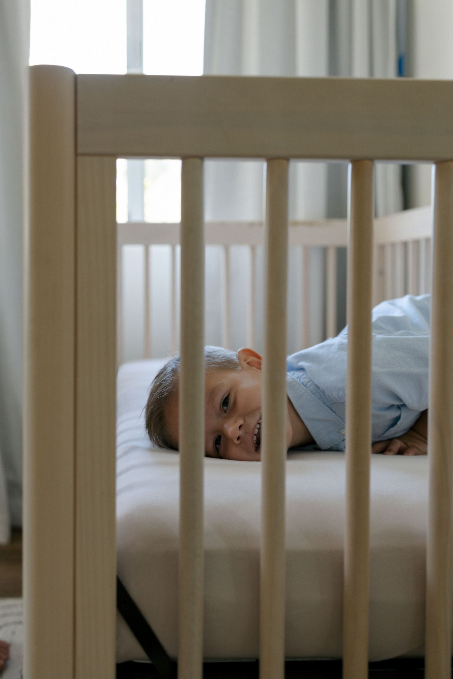 Young child lying down smiling in a white wooden crib, wearing a light blue shirt in a bright nursery room.
